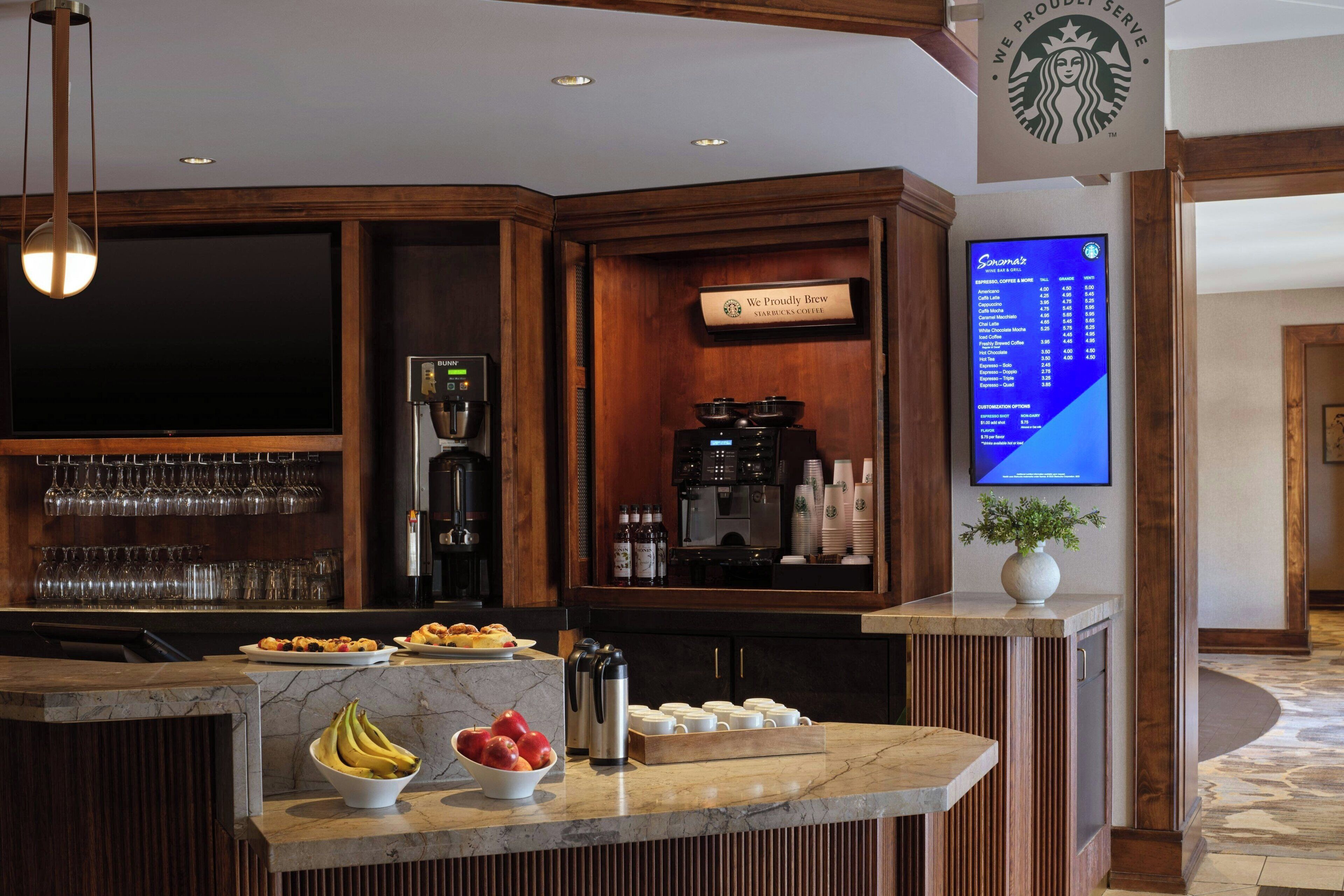 A hotel bar area with a coffee machine, a menu board, and various food items on the counter.