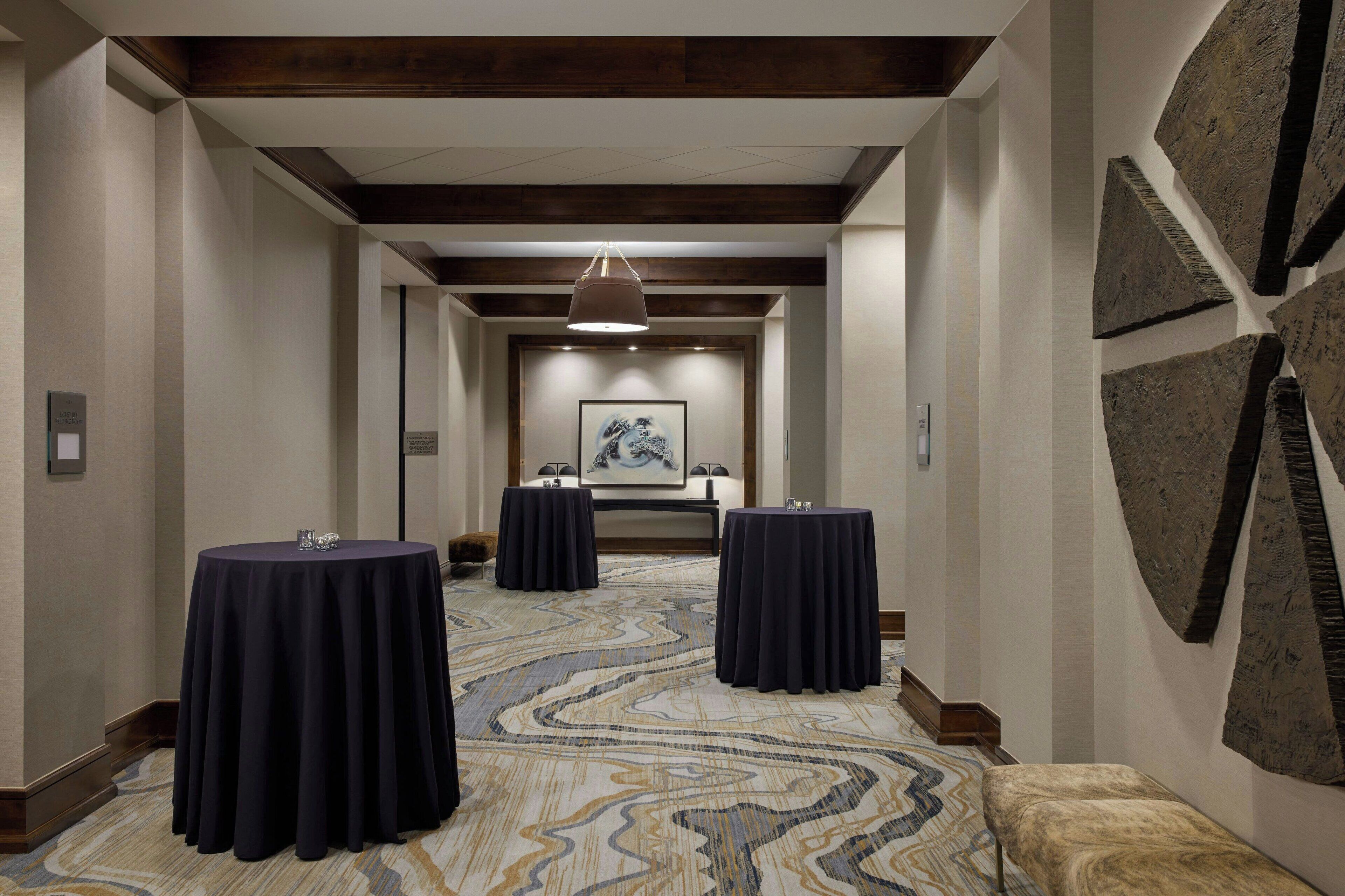 A hotel corridor with three round tables, a patterned carpet, and a framed artwork on the wall.