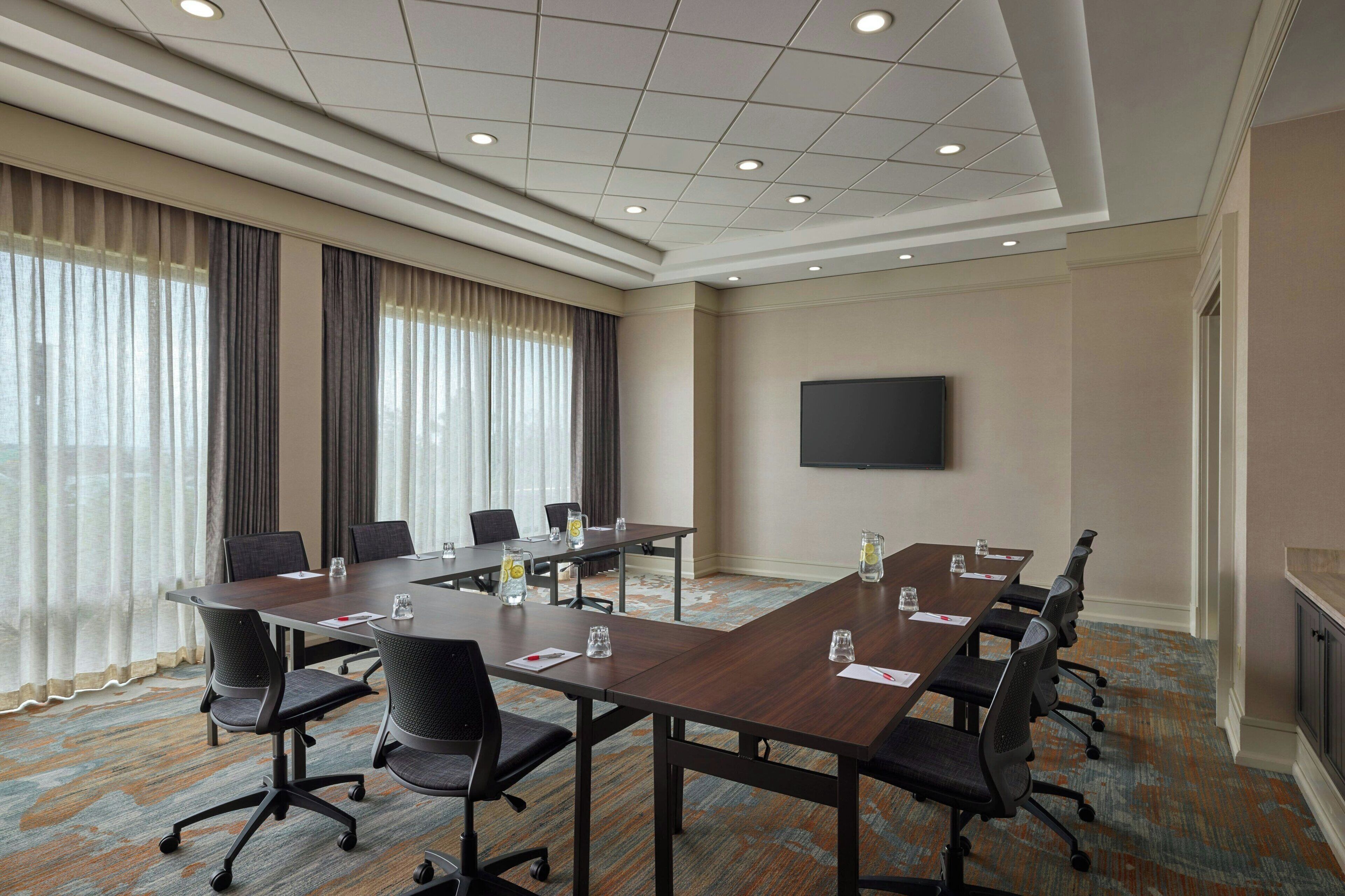 A conference room with a long table, chairs, a flat-screen TV, and a large window with curtains.