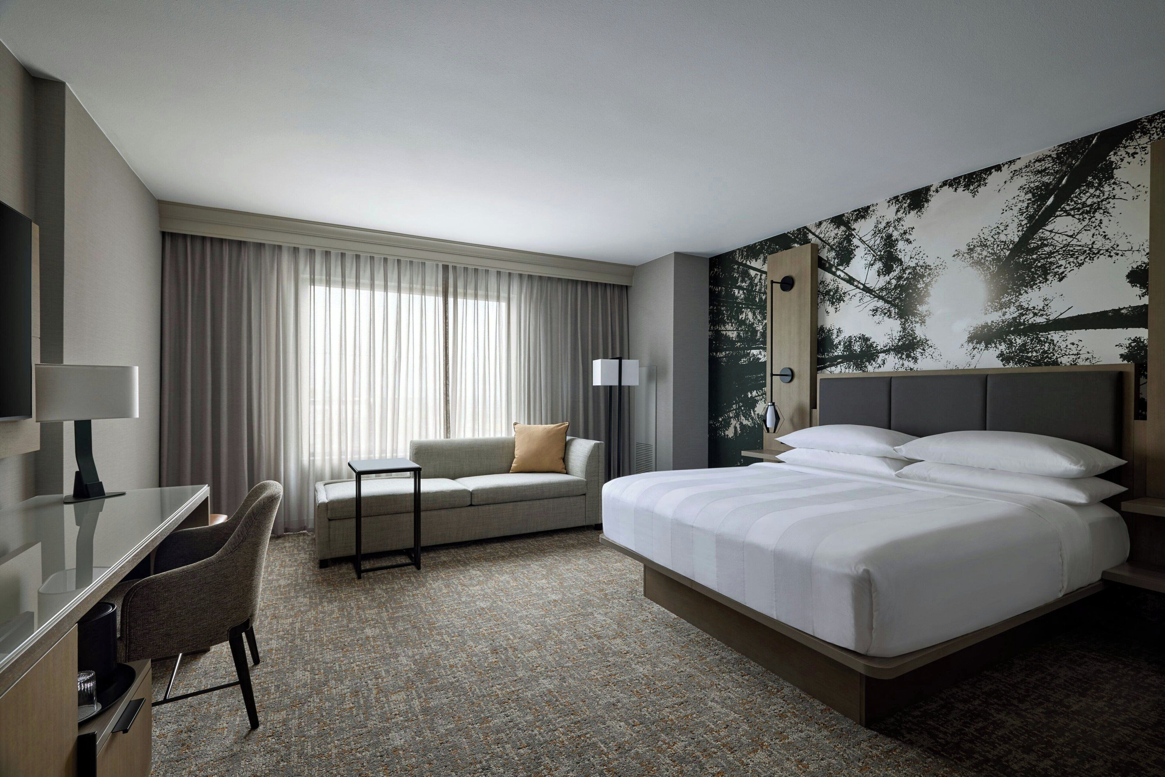 Une chambre d’hôtel avec un lit, un bureau, une chaise, un canapé et une grande fenêtre avec des rideaux.