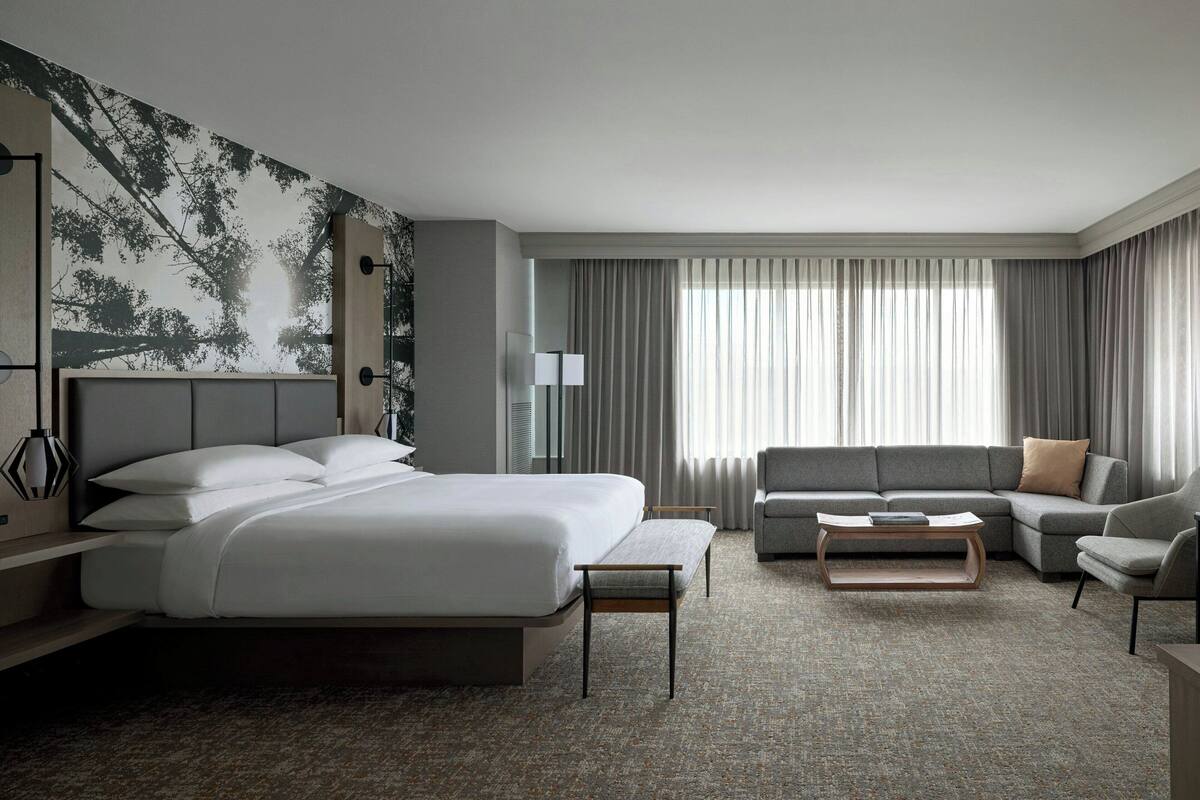 Habitación de hotel moderna con una cama grande, un sofá, una silla y una mesita de café. Cuenta con un amplio ventanal con cortinas y una pared con un mural de temática natural.