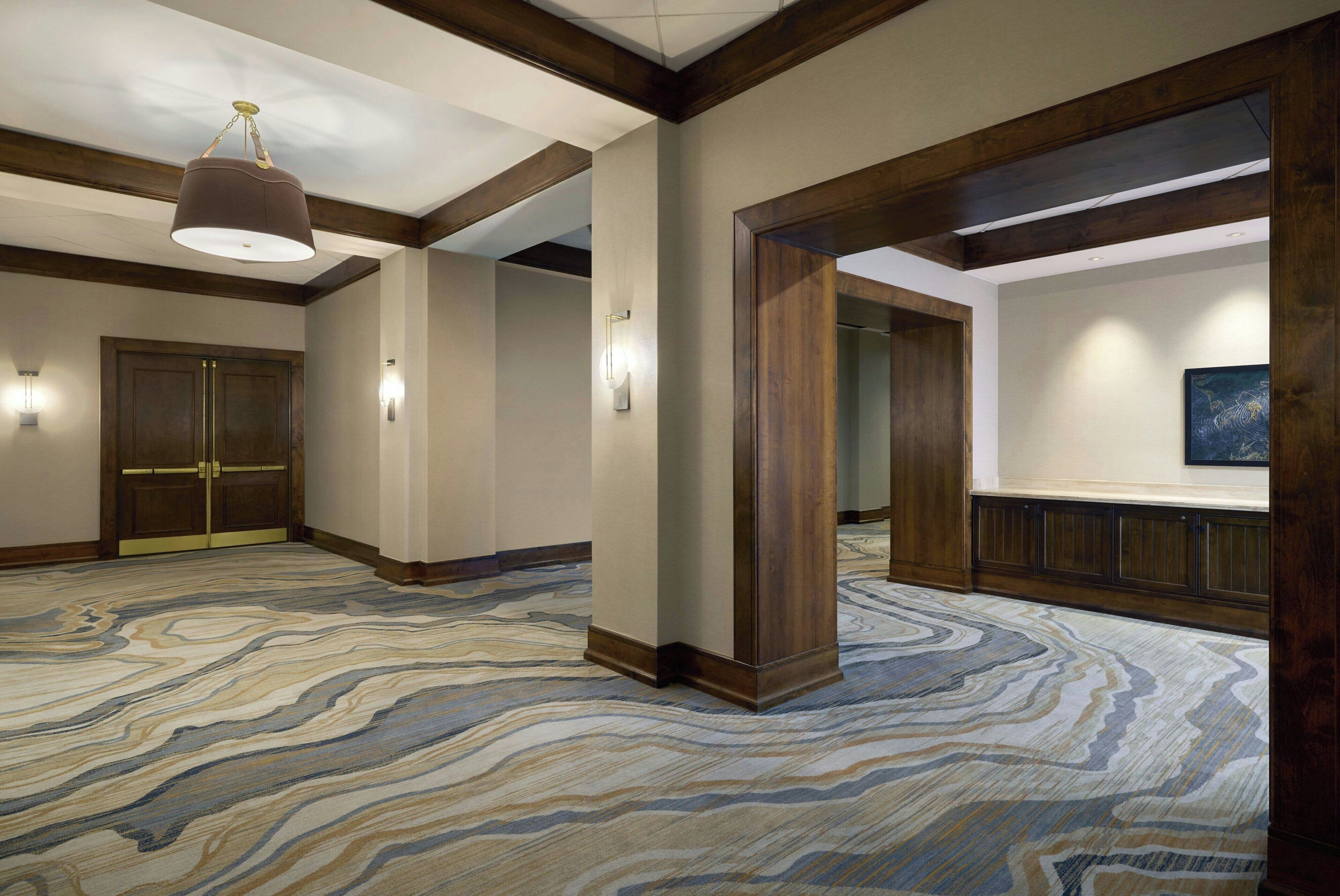 A spacious hotel lobby with a marble-patterned carpet, wooden columns, and a large chandelier.