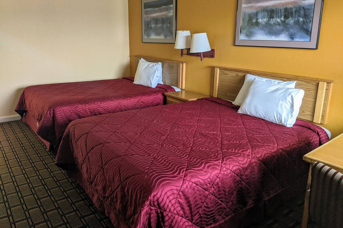 A hotel room with two beds, each with a red quilt, a wooden headboard, and a nightstand with a lamp.