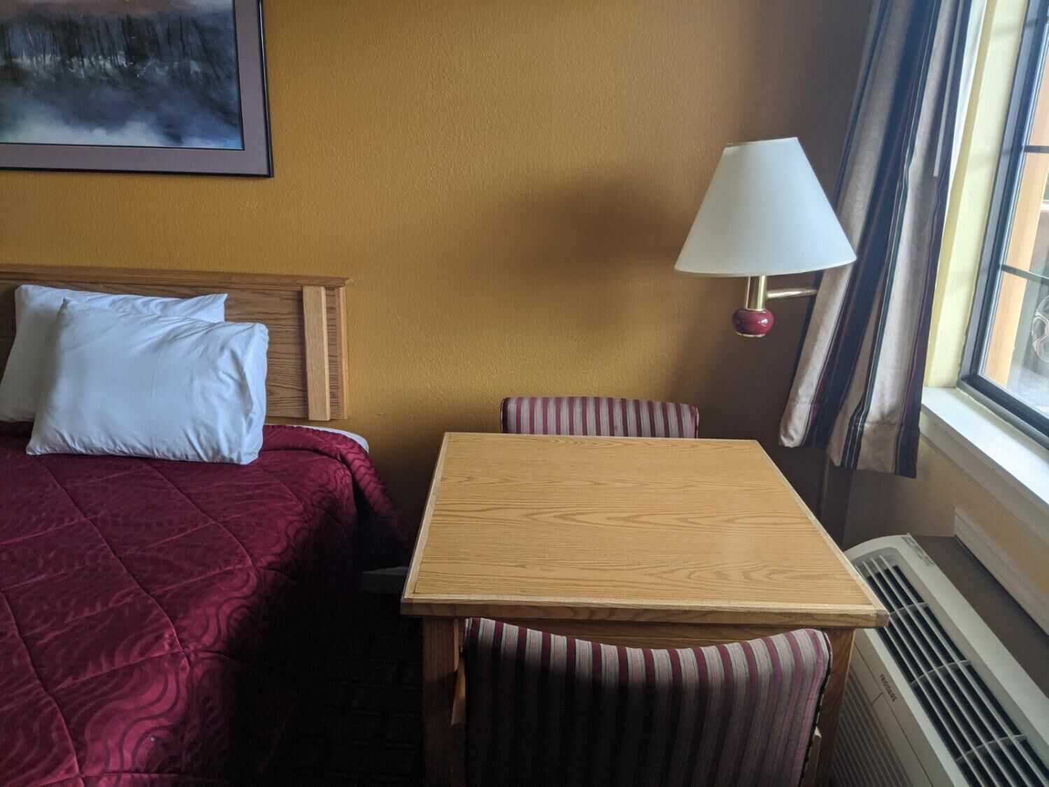 A hotel room with a bed, a table, and a chair. There is a window with curtains, a wall-mounted lamp, and a painting above the bed.