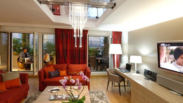 A modern living room with a high ceiling, a chandelier, a TV, and a seating area with red sofas and a coffee table. - Résidence Alma Marceau (Paris)