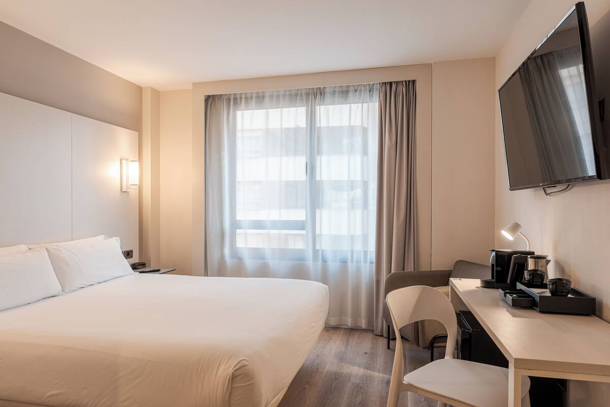 Habitación de hotel con cama, escritorio con lámpara, televisión y ventana con cortinas.