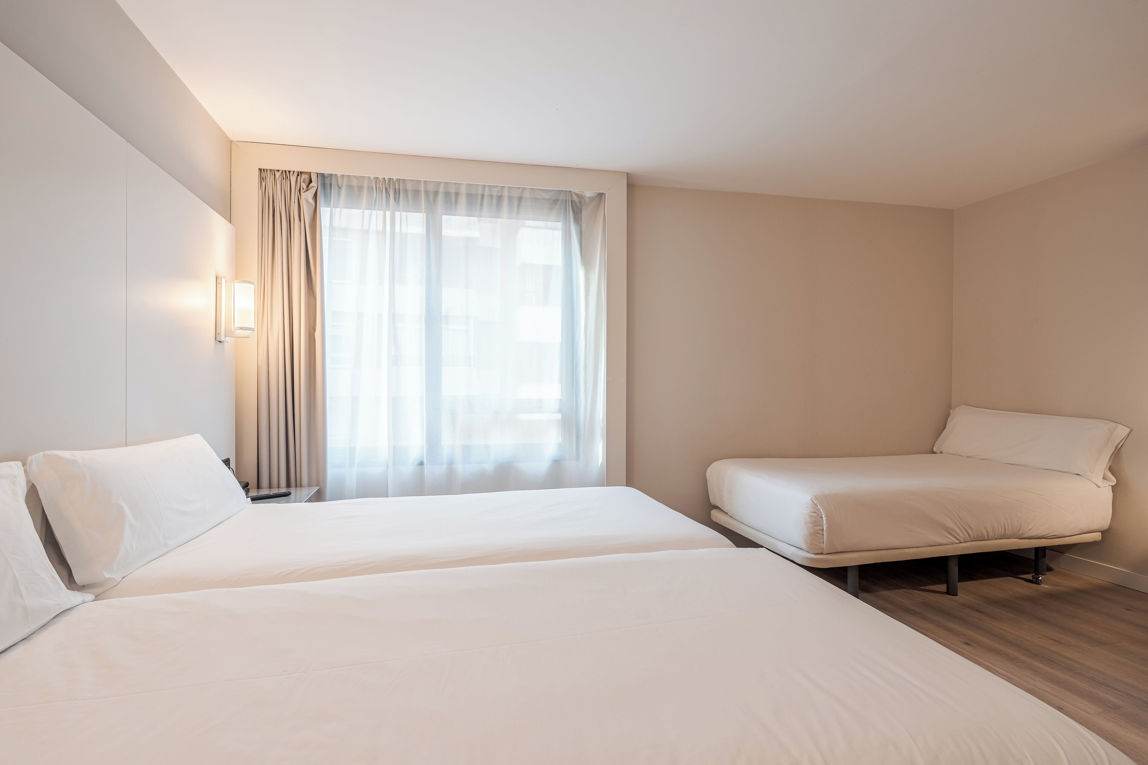 A hotel room with two beds, a window with curtains, and a bedside table with a lamp.