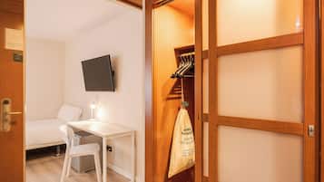 A room with a bed, a desk, a chair, a TV mounted on the wall, and a wooden wardrobe with a hanging bag.