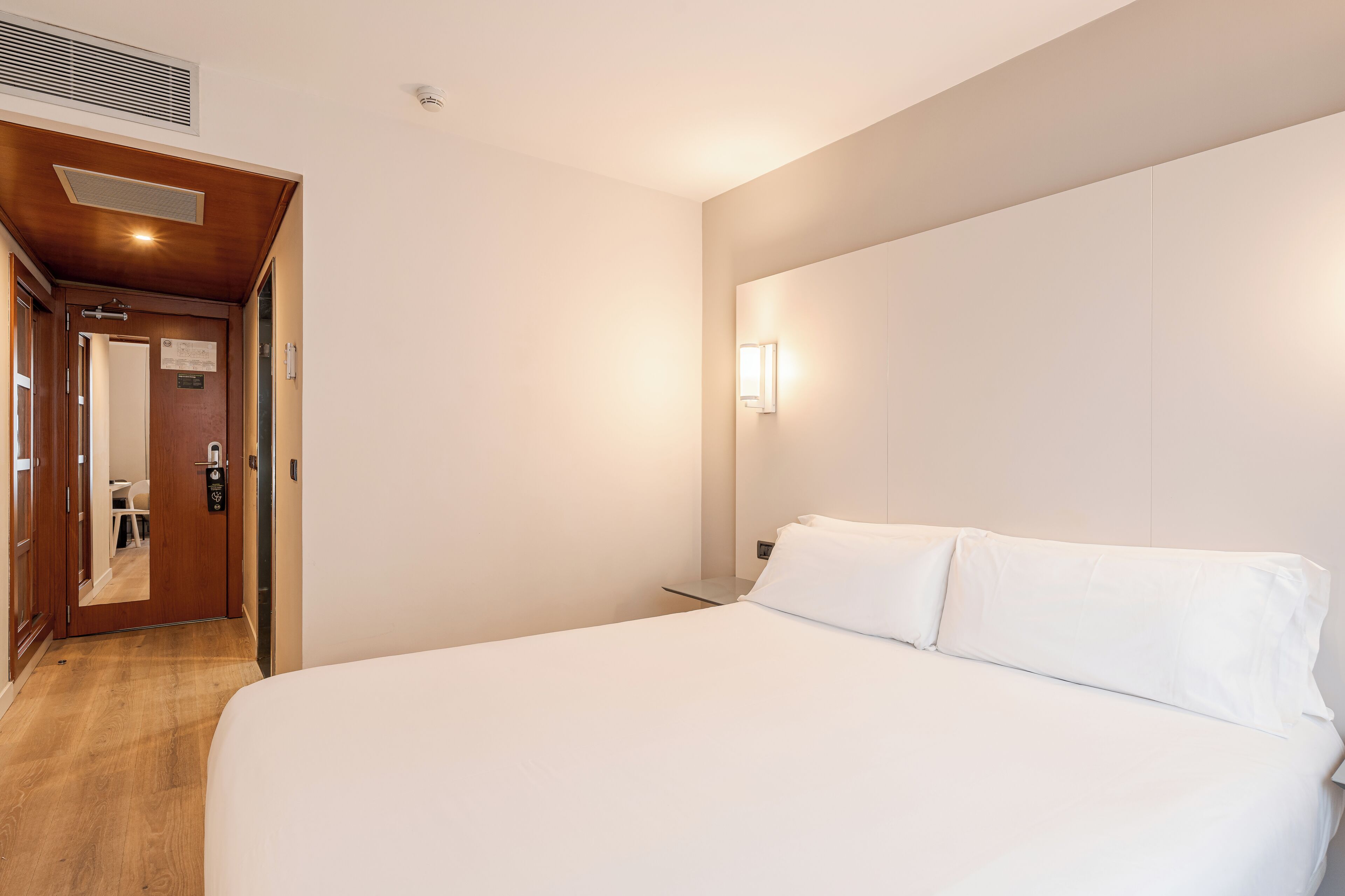 A hotel room with a wooden floor, a bed with white linens, a bedside table, a wall-mounted light, and a door leading to a bathroom.