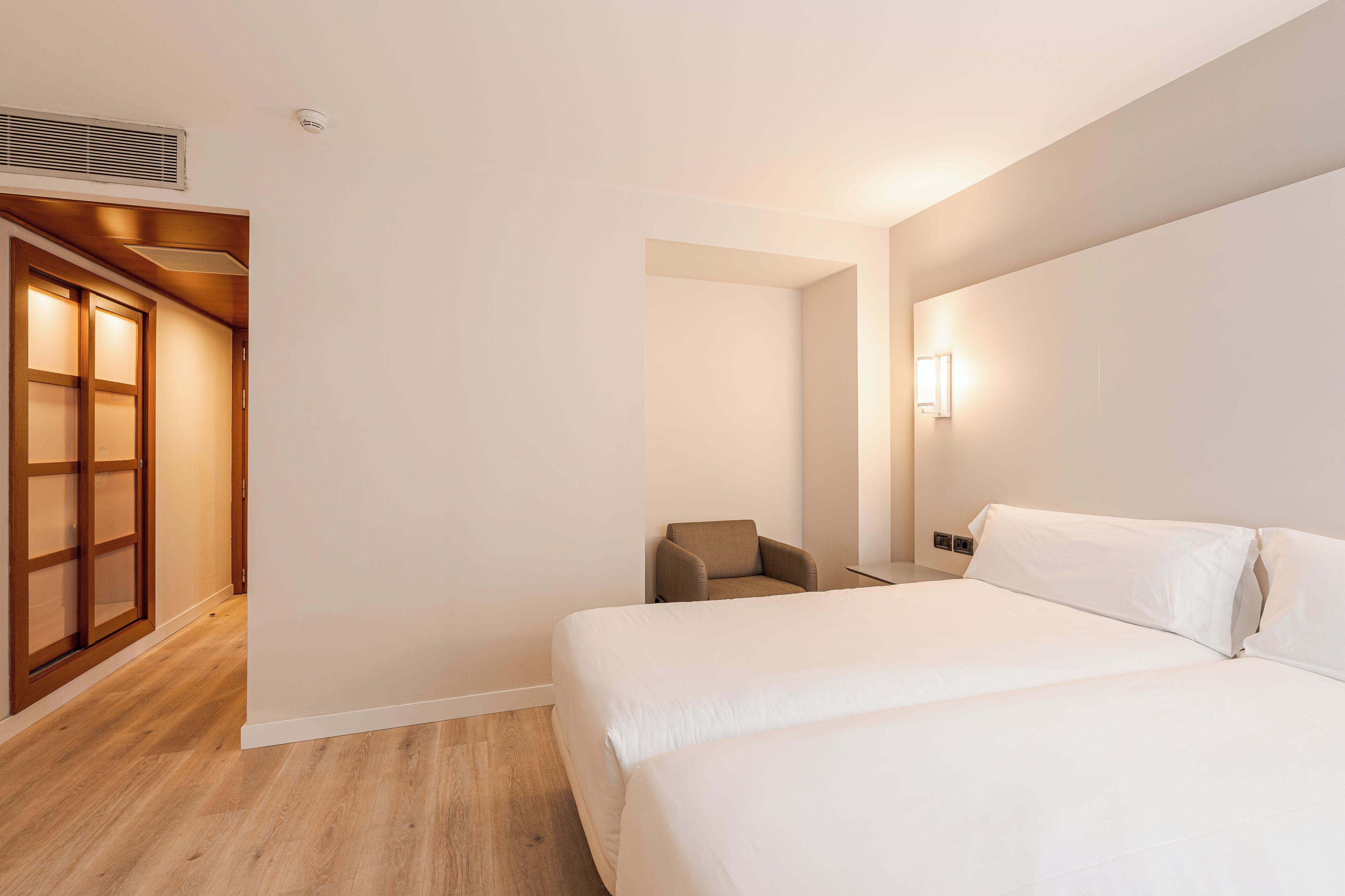 Una habitación de hotel con una cama, una silla, una lámpara de pared y una puerta de madera.