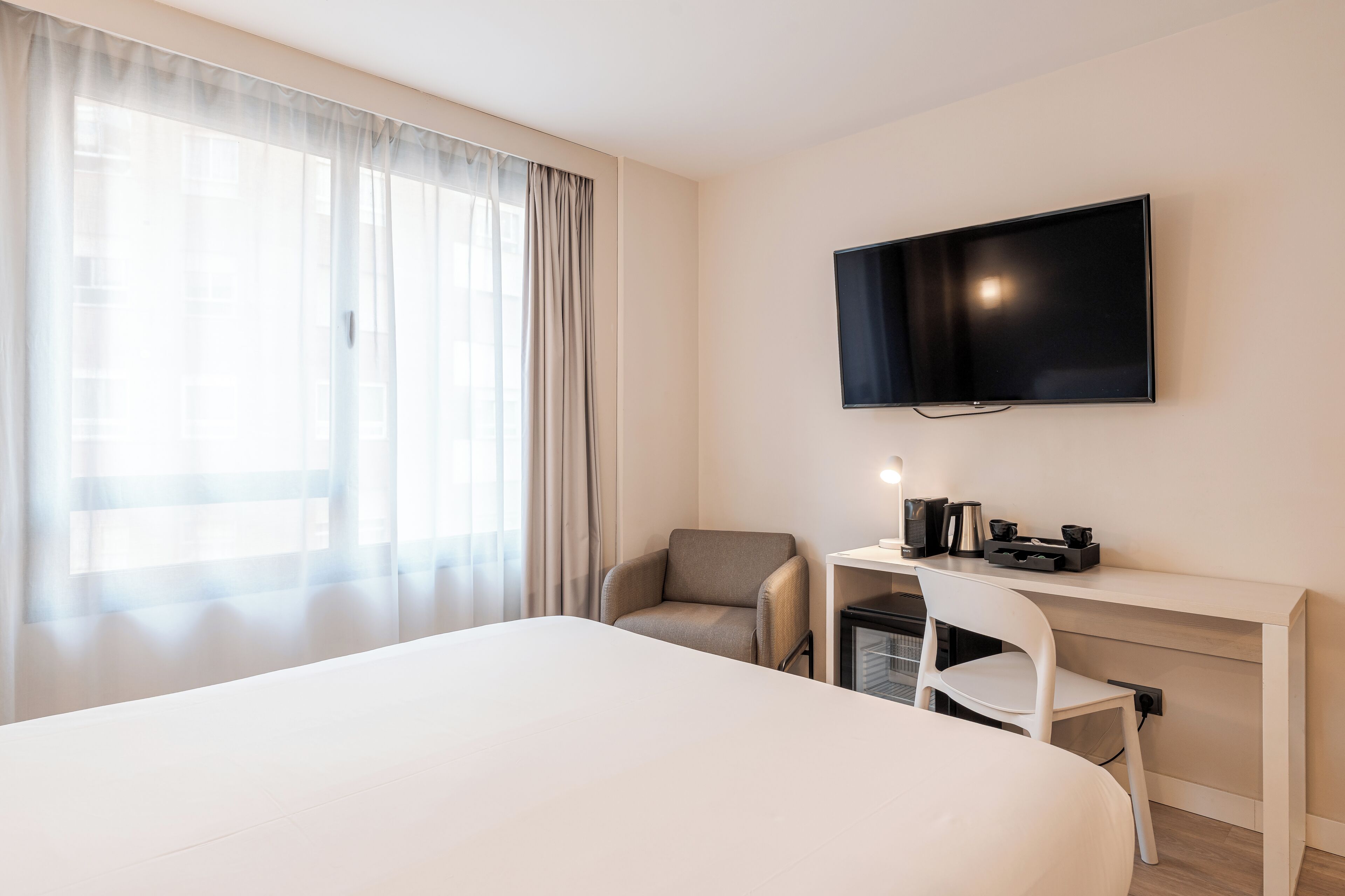 A hotel room with a bed, a desk with a chair, a television, and a window with curtains.