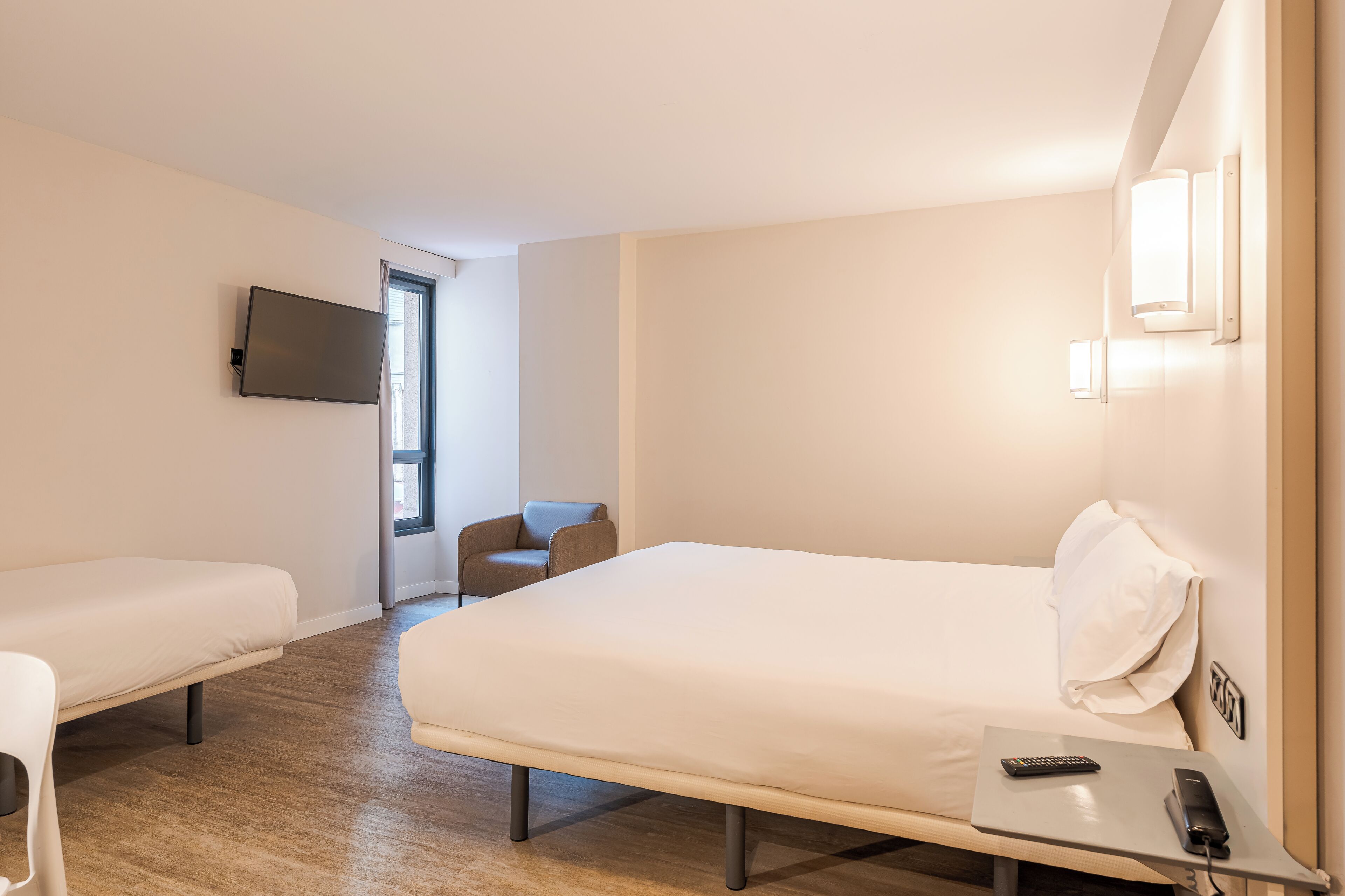 A hotel room with a large bed, a smaller bed, a desk with a remote control, a chair, and a TV mounted on the wall.