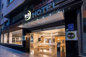 A hotel entrance with a glass facade, a reception desk, and a visible interior seating area.