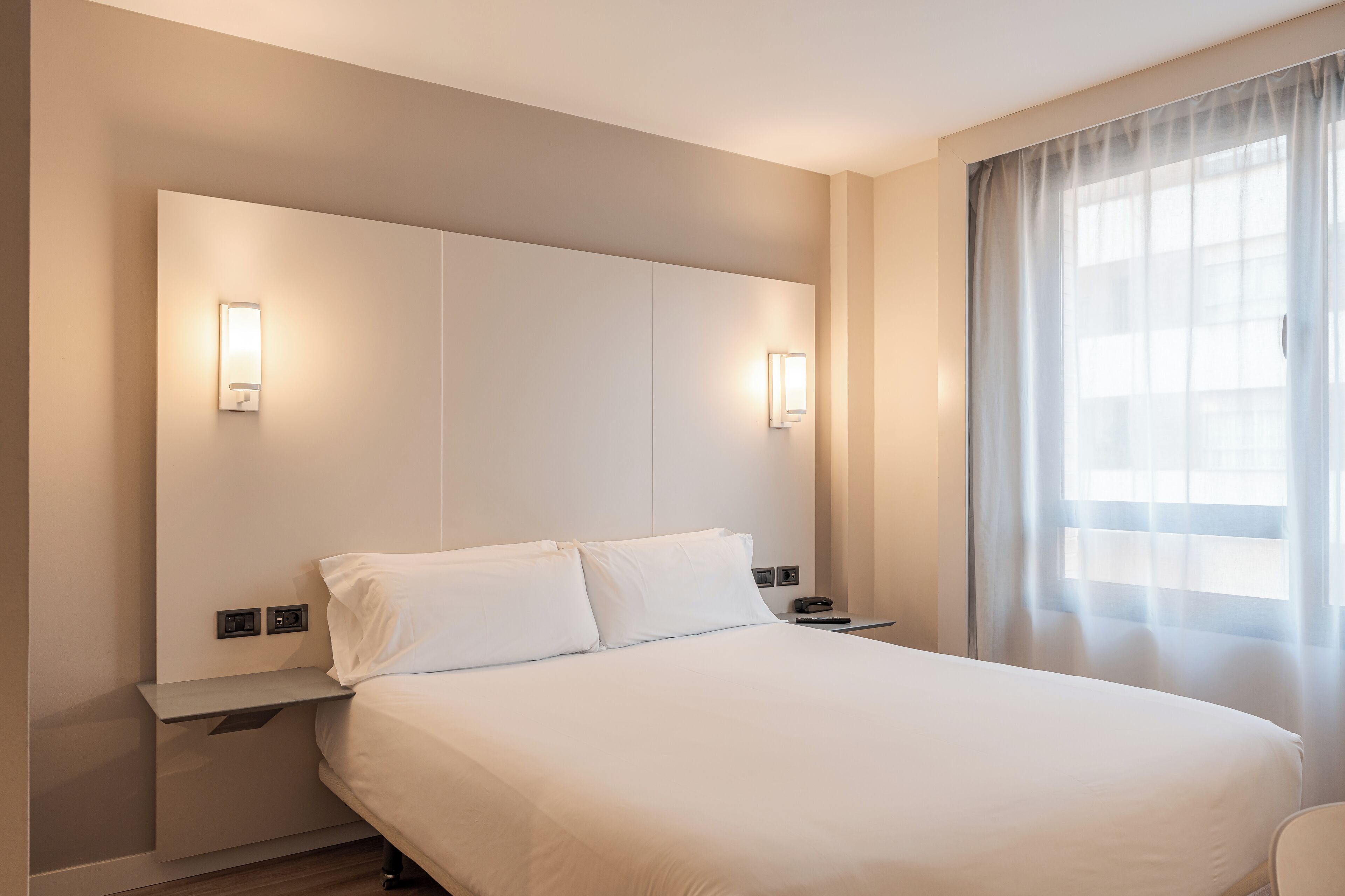 Una habitación de hotel moderna con una cama grande, dos mesitas de noche, un cabecero y un ventanal con cortinas.