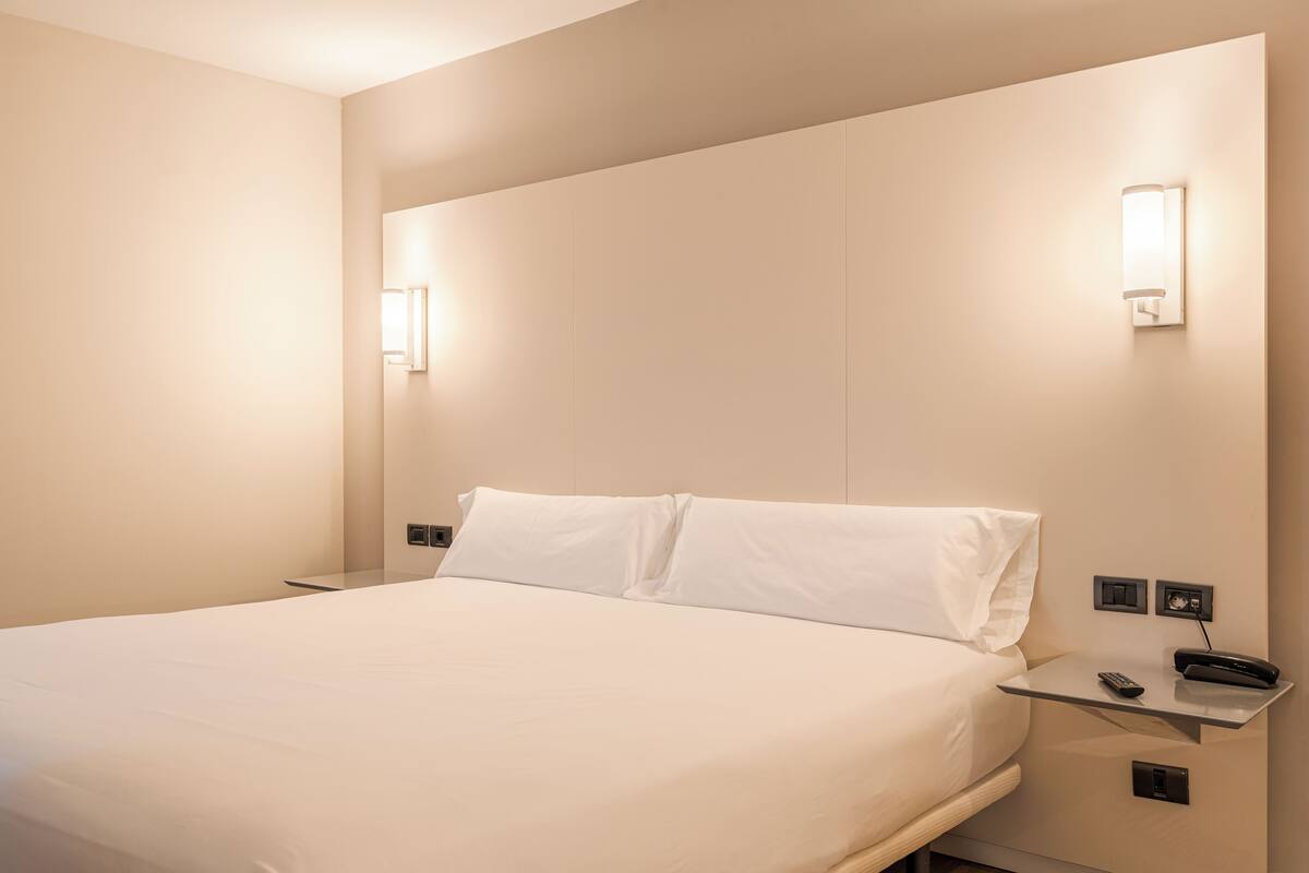 Una habitación de hotel con cama, mesitas de noche, teléfono y una lámpara fijada a la pared.