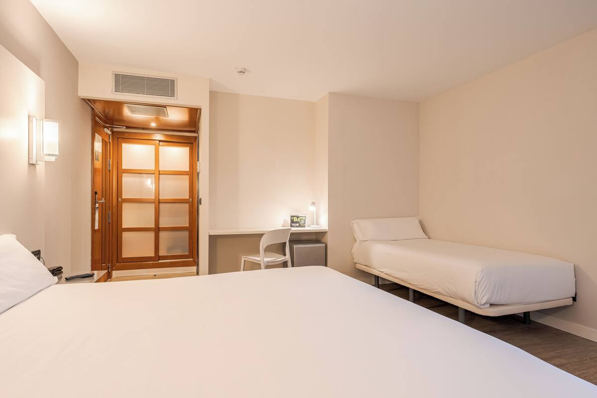 Una habitación de hotel con una cama grande, un escritorio con silla, un armario con espejo y una mesita de noche.