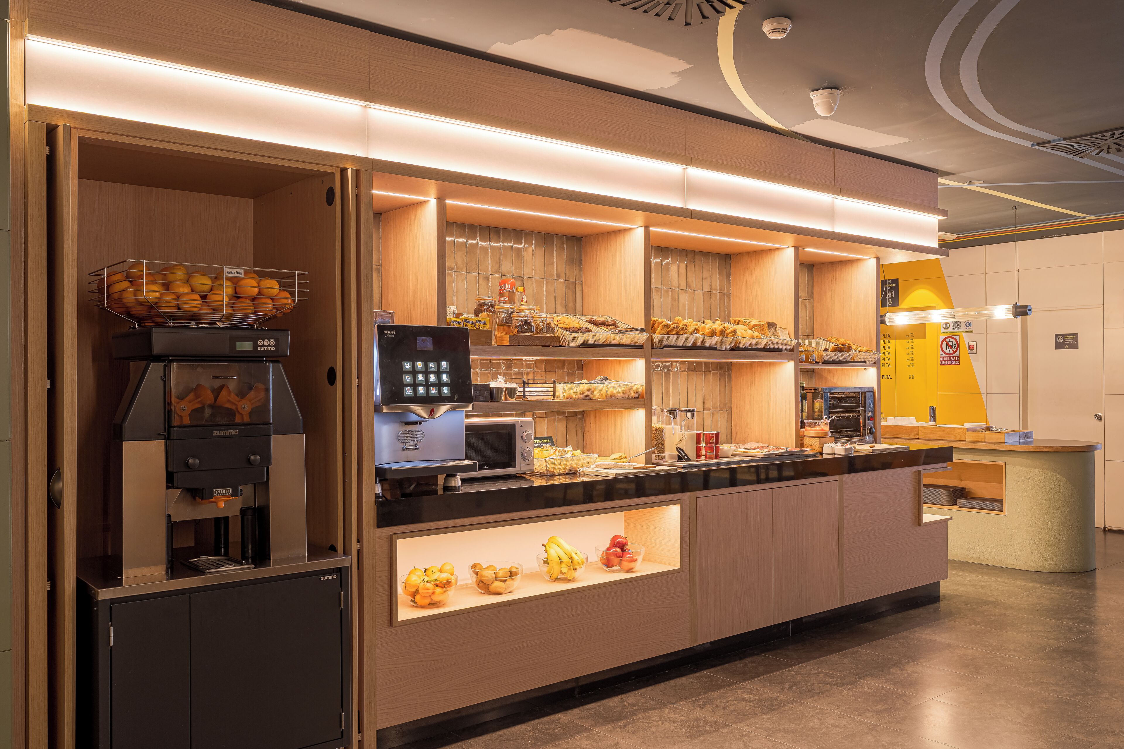 A self-service breakfast station with a coffee machine, fruit display, and bread.