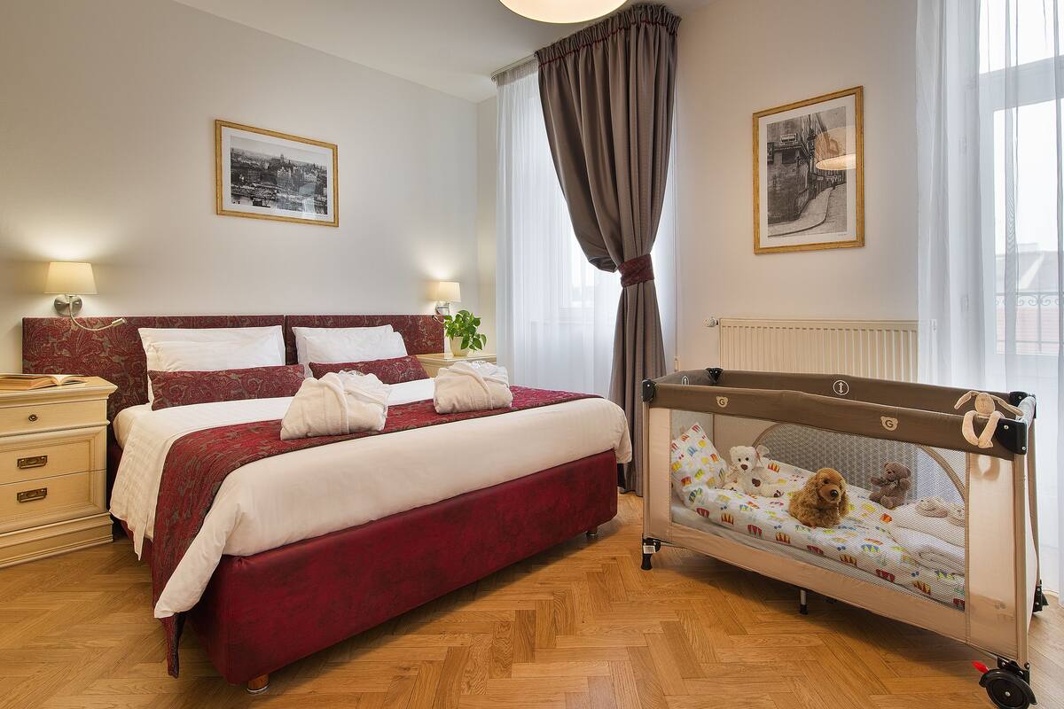 A hotel room with a large bed, a bedside table with a lamp, a crib with a teddy bear, and two framed pictures on the wall.