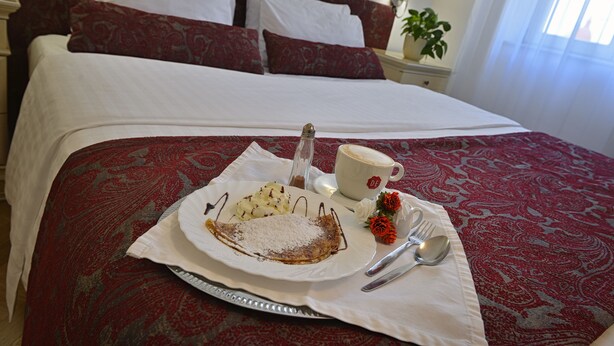 A neatly made bed with a red and white patterned bedspread, a plate of food including a crepe, a cup of coffee, and cutlery on a tray. - Hotel Suite Home Prague (Prague)