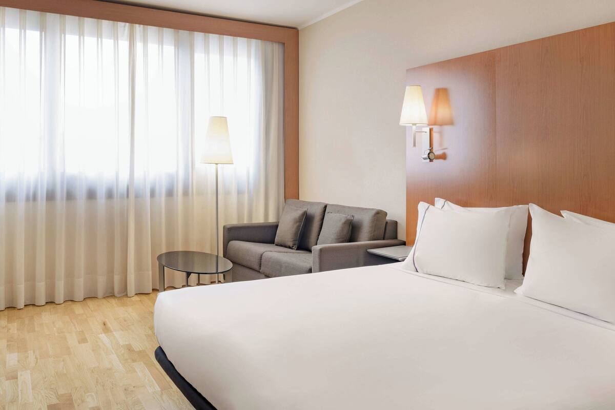 A hotel room with a large bed, a grey sofa, a small table, and two wall-mounted lamps.