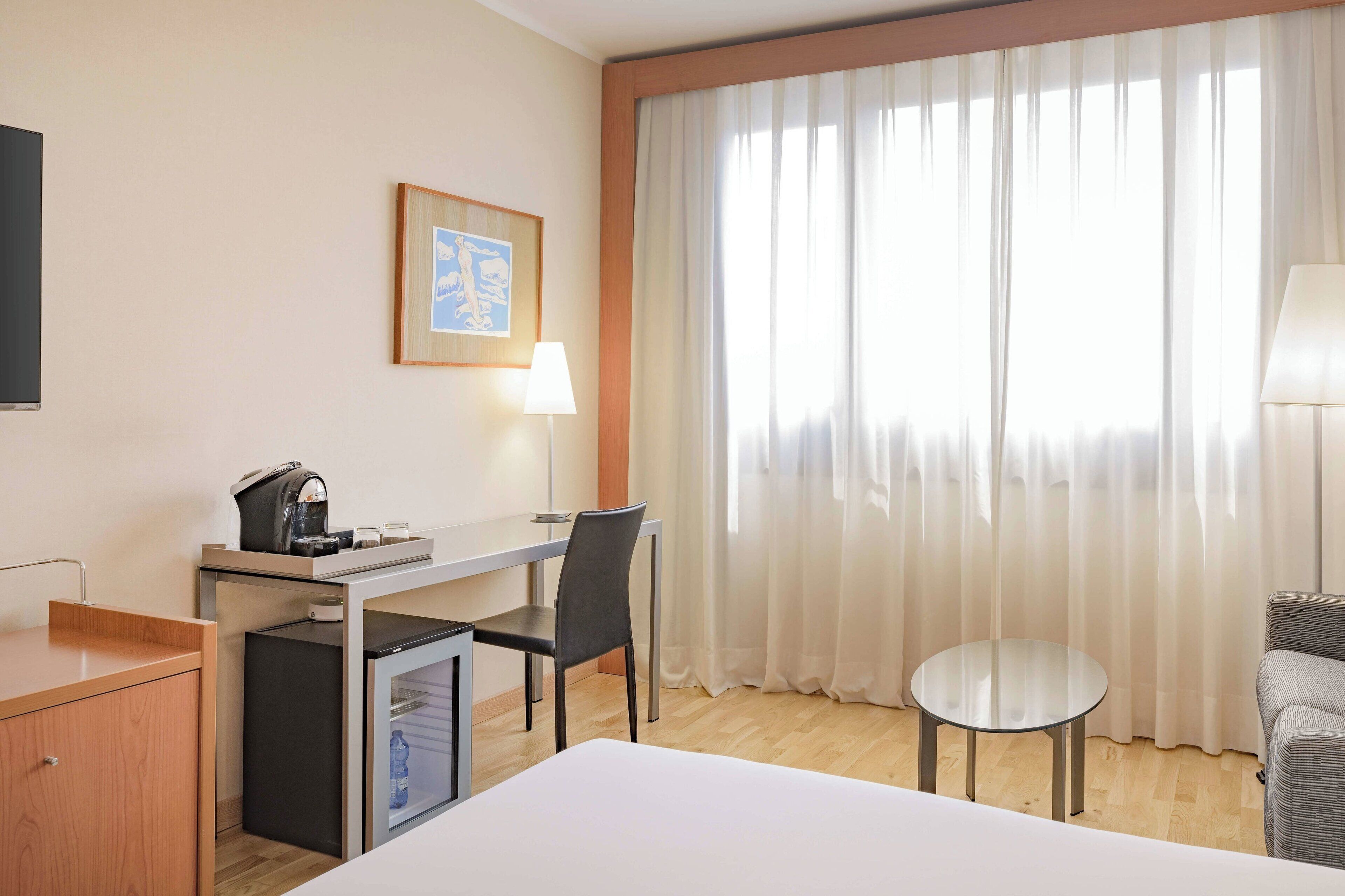 A hotel room with a bed, a desk with a chair, a small fridge, a coffee machine, a lamp, and a window with sheer curtains.