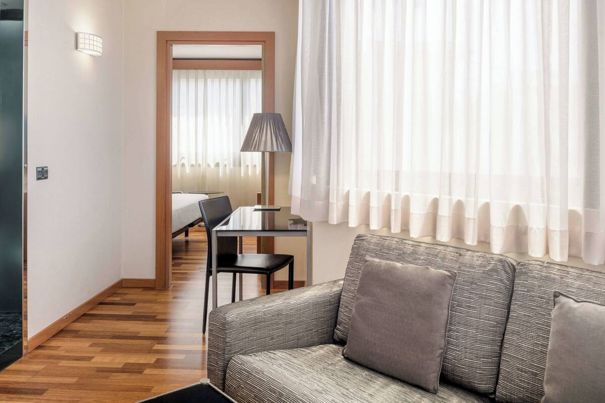 A hotel room with a sofa, a dining table with chairs, a lamp, and a bed with a wooden headboard.