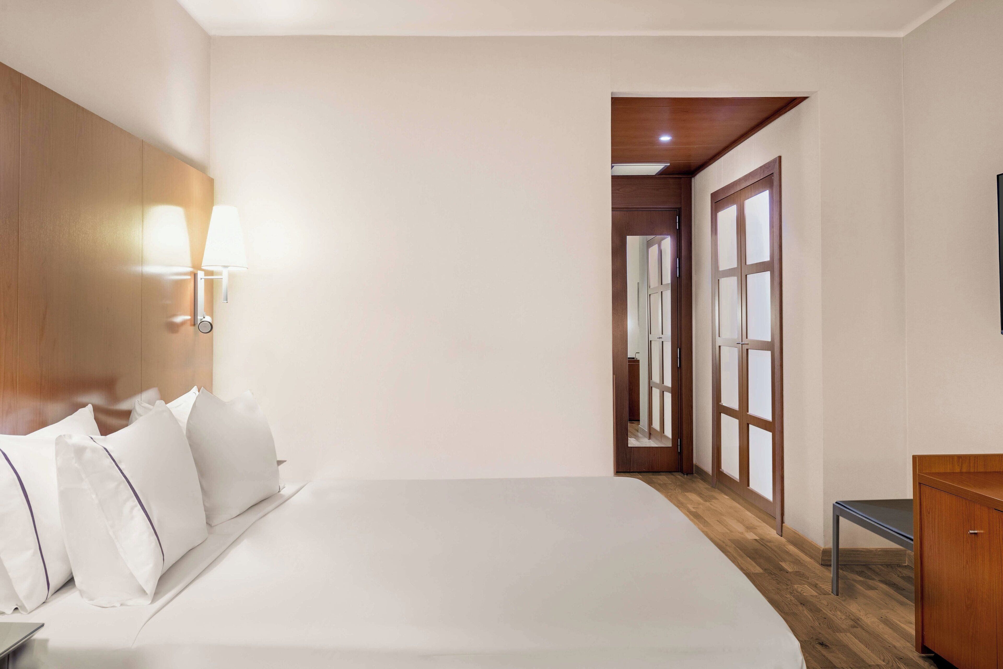 A hotel room with a large bed, a wooden headboard, a nightstand, a desk, and a wardrobe.