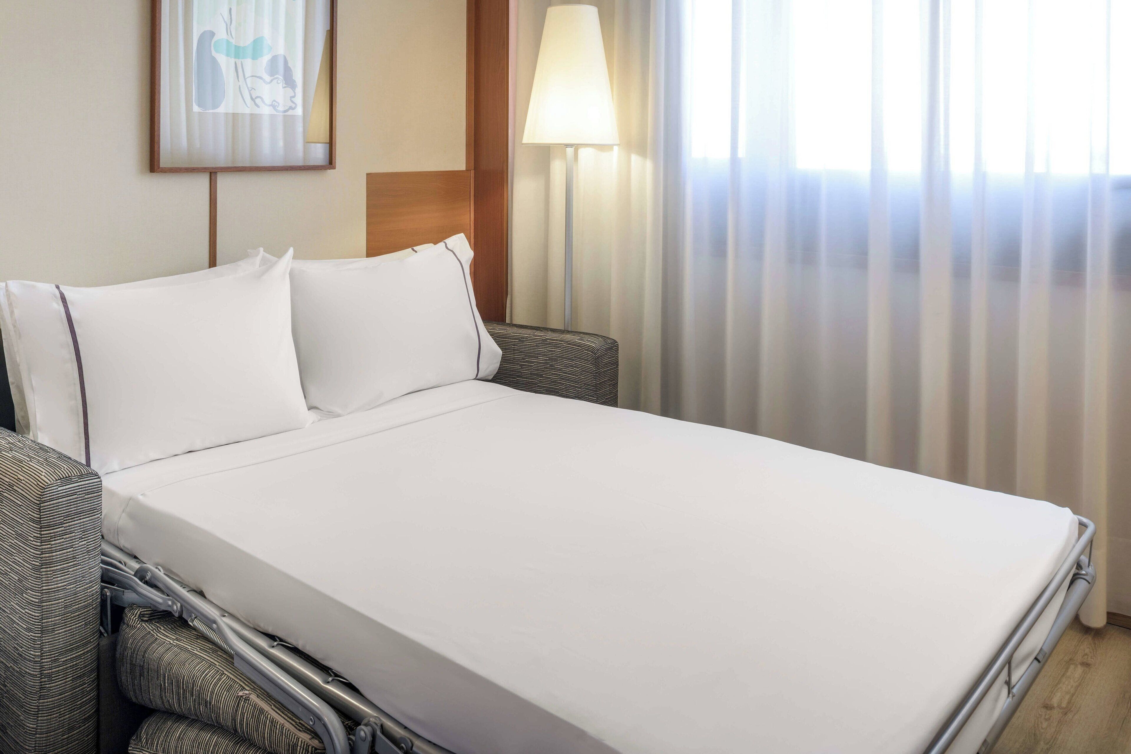 A neatly made bed with white linens, a grey upholstered headboard, and a bedside lamp.