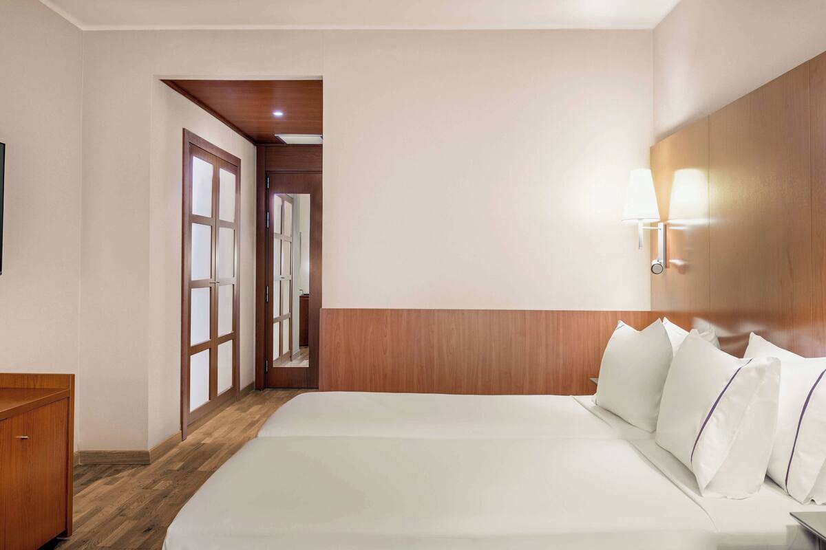 A hotel room with a wooden headboard, a bed with white bedding, a nightstand, and a wall-mounted lamp.