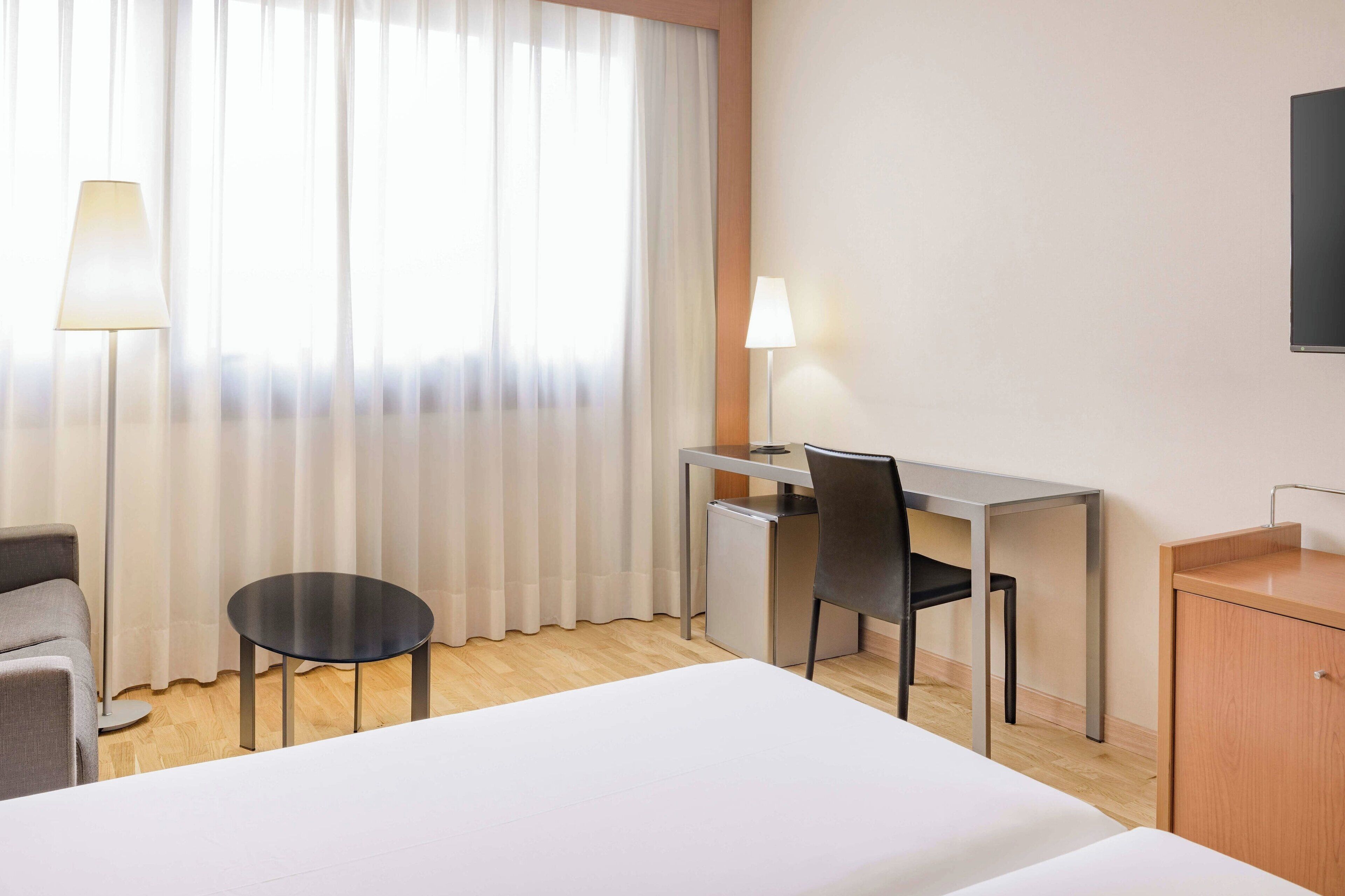 A hotel room with a bed, a desk, a chair, a small table, a lamp, and a TV.