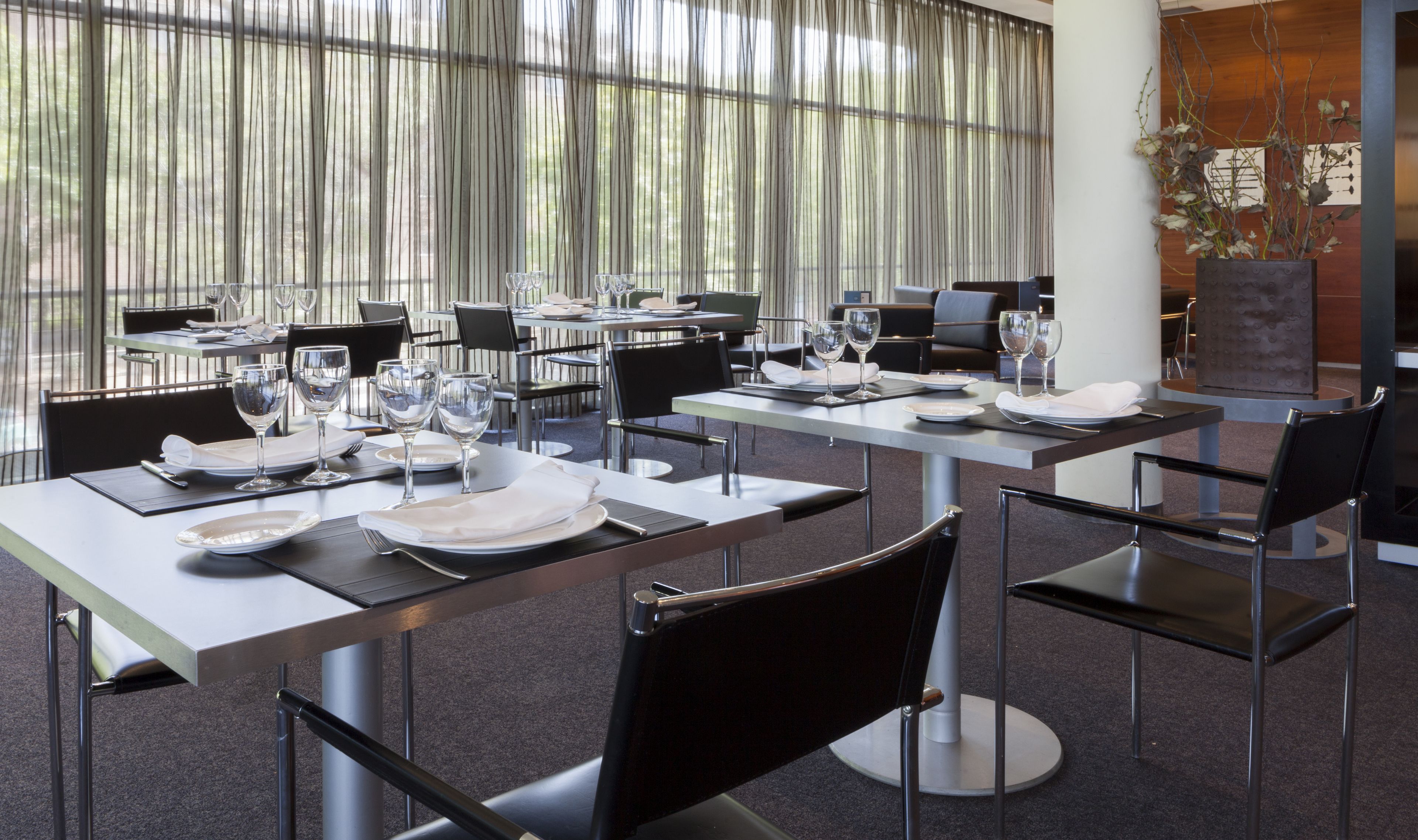 Un restaurante con mesas preparadas para comer, con sillas modernas y un amplio ventanal con cortinas translúcidas.
