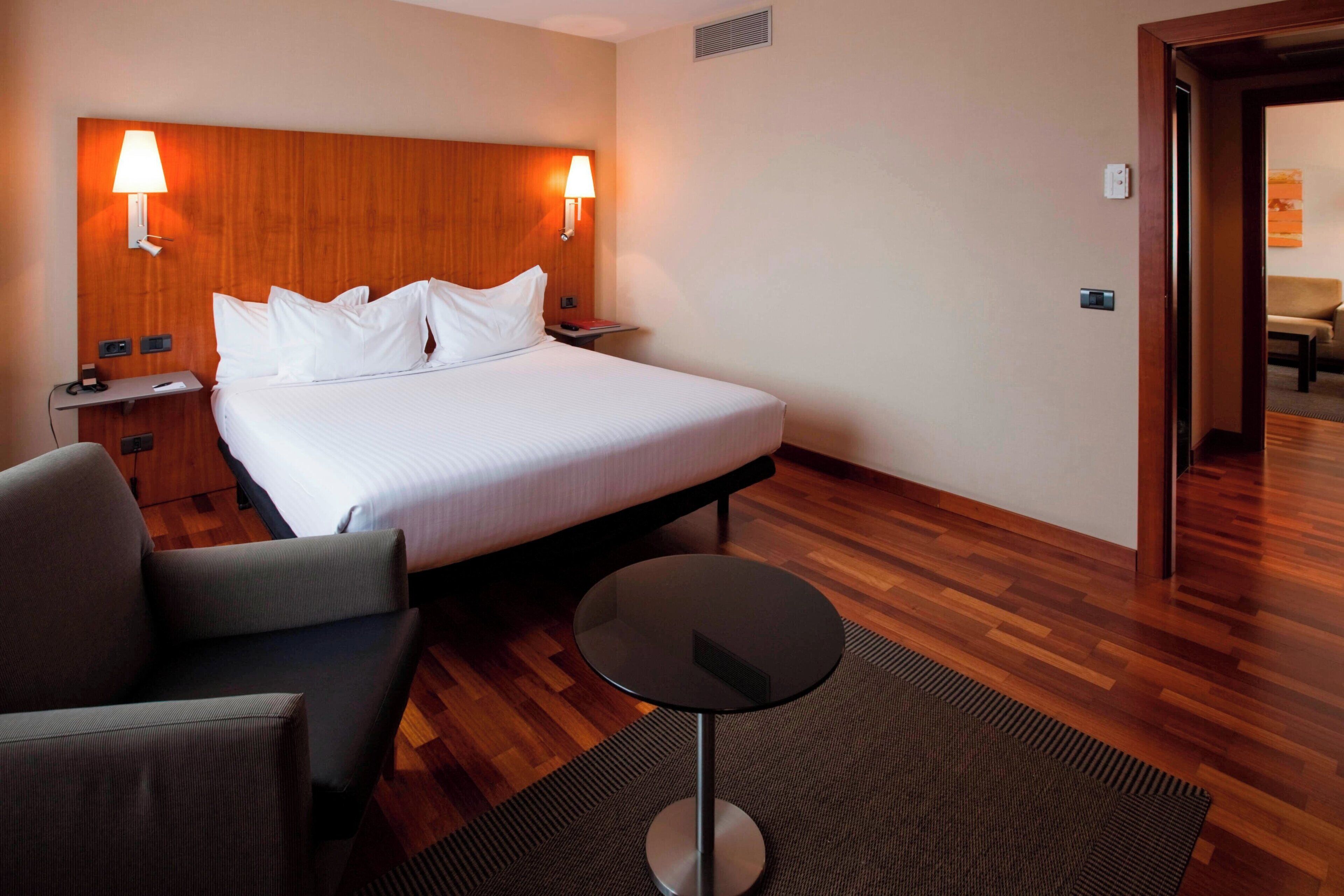 Habitación de hotel con cama, dos lámparas de noche, una mesita redonda, una silla y suelo de madera.