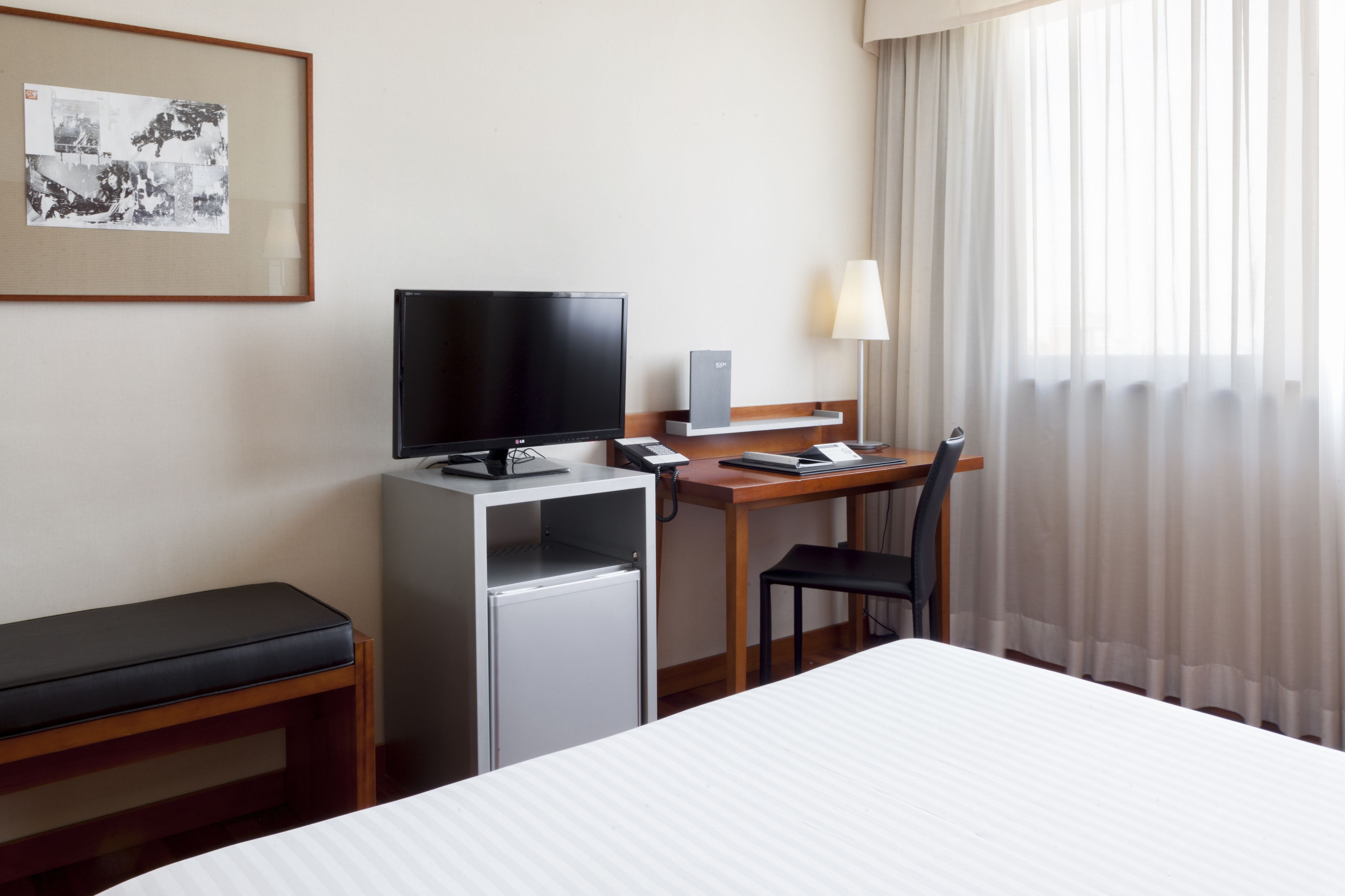 Habitación de hotel con cama, escritorio con computadora, televisor, refrigerador pequeño, silla y ventana con cortinas.