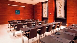 Uma sala de conferências com mesas compridas e cadeiras, uma placa da AC Hotels Marriott e duas grandes obras de arte emolduradas nas paredes.