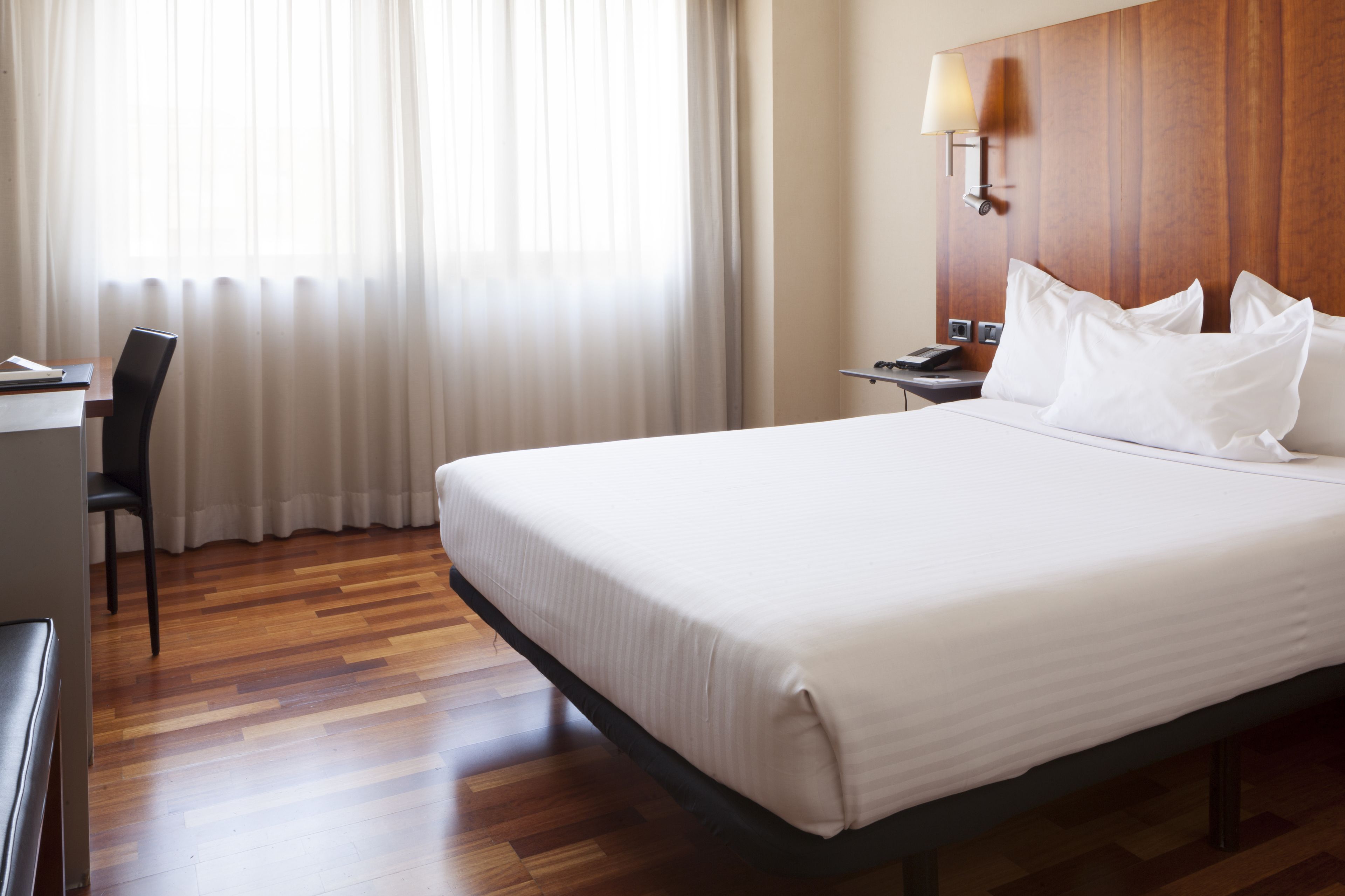 A hotel room with a large bed, a desk, a chair, and a nightstand with a lamp.