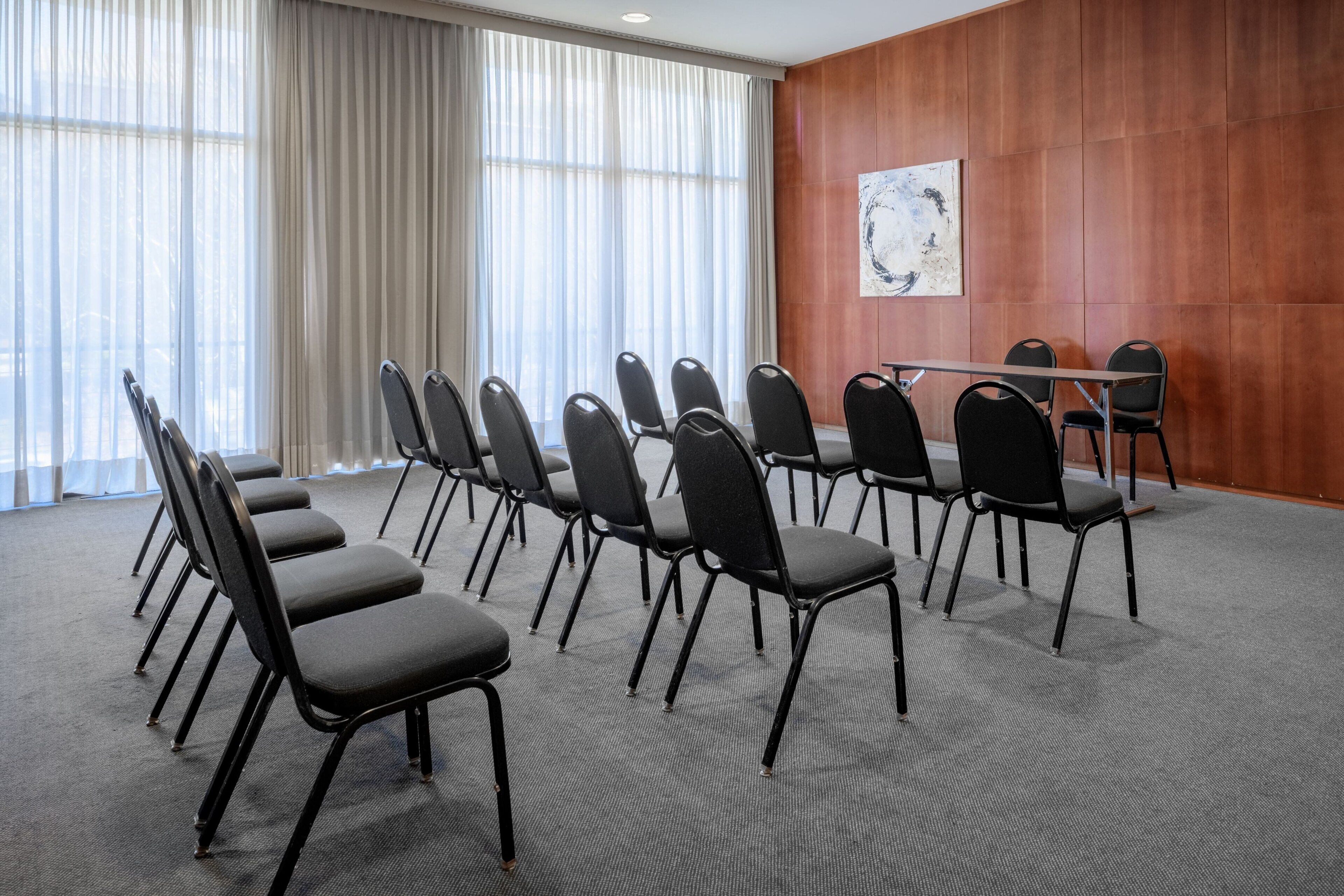 Ein Konferenzraum mit Reihen schwarzer Stühle, einem langen Tisch und einem großen Fenster mit transparenten Vorhängen.