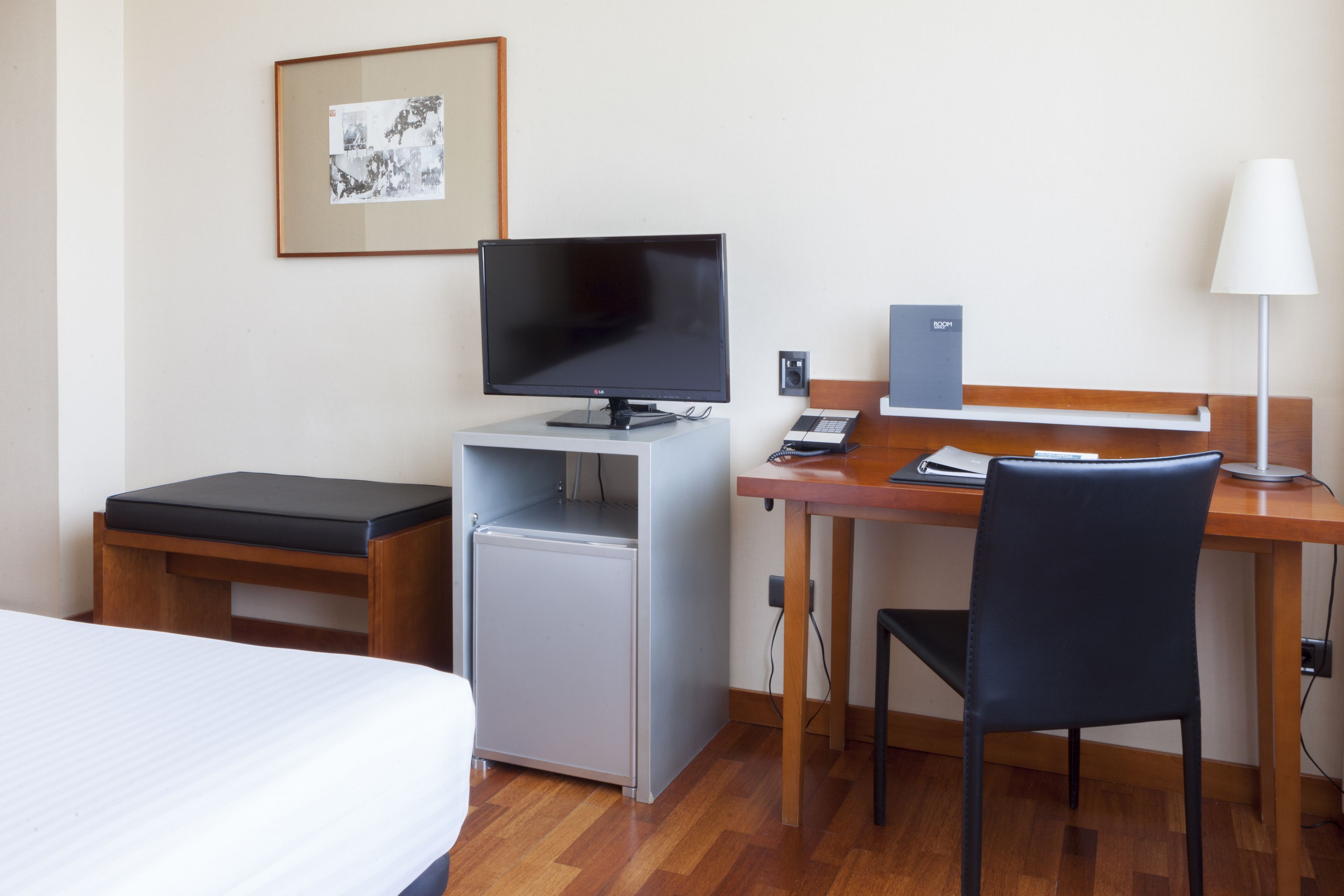 A compact hotel room with a bed, a desk with a computer, a chair, a small refrigerator, and a lamp.