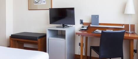 A compact hotel room with a bed, a desk with a computer, a chair, a small refrigerator, and a lamp.