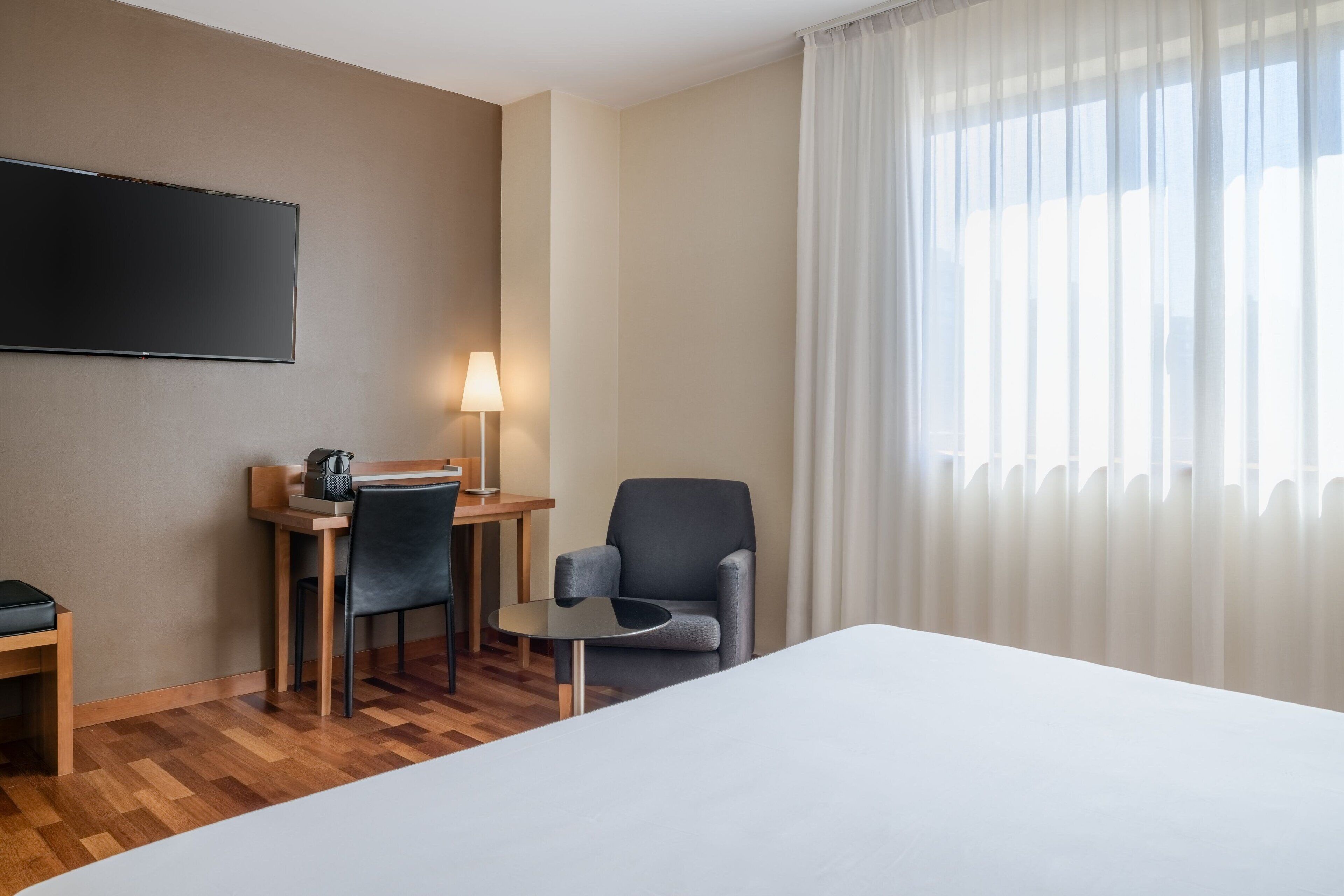 A hotel room with a bed, a desk with a chair, a lamp, and a TV on the wall.