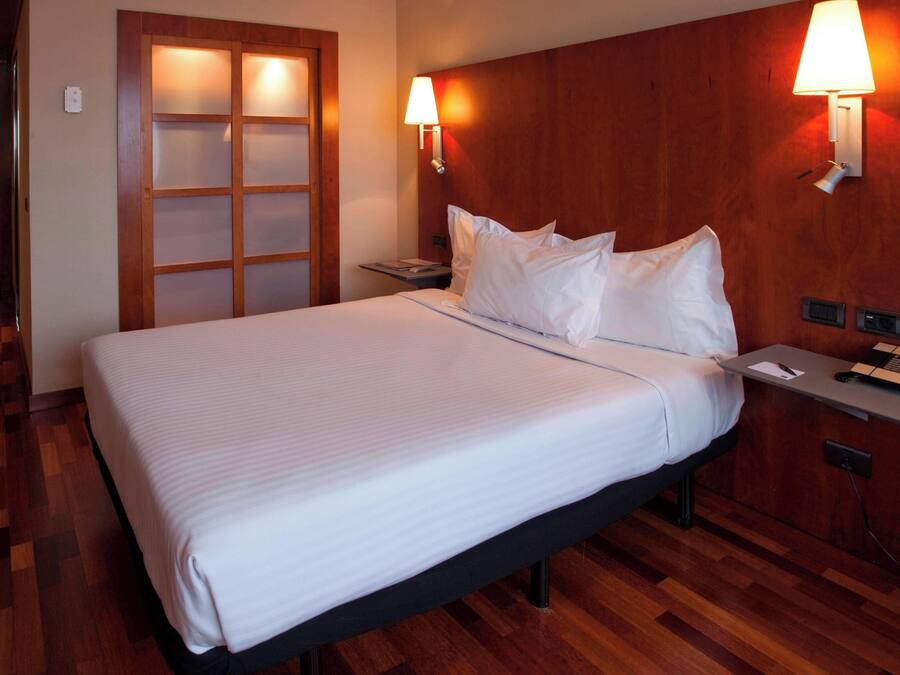 A hotel room with a large bed, wooden headboard, two bedside lamps, a nightstand with a phone, and a wooden floor.