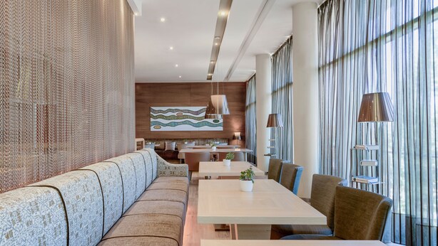 Uma área de refeições moderna com um longo banco, mesas e cadeiras, e uma cozinha ao fundo.