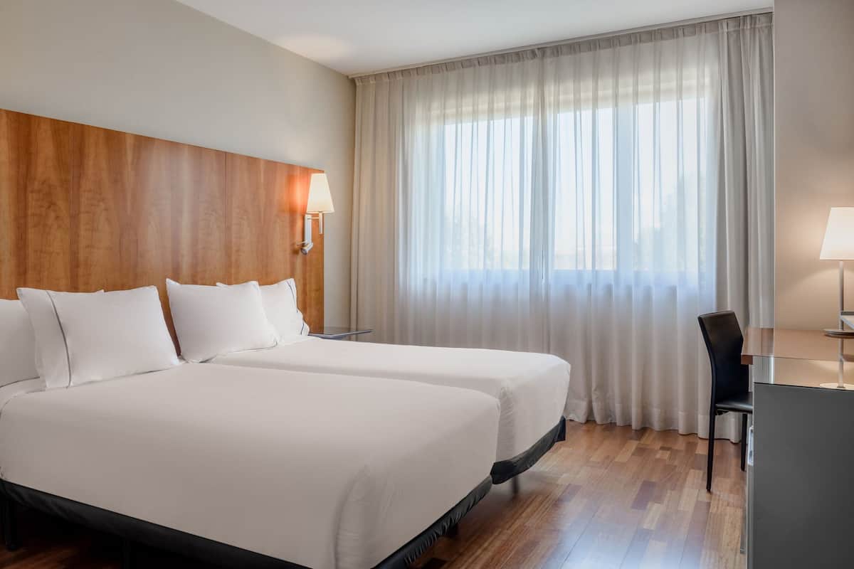 Een hotelkamer met twee bedden, een houten hoofdbord, een bureau met stoel, en een groot raam met dunne gordijnen.
