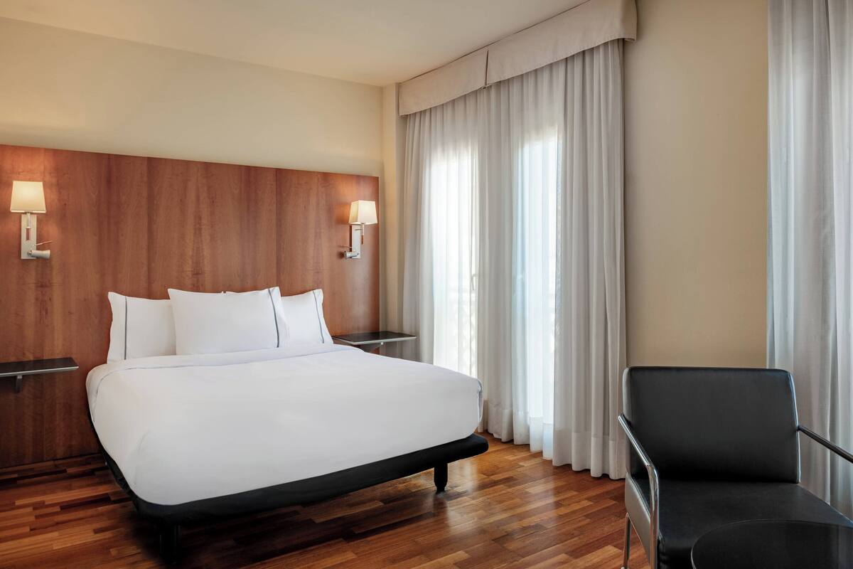 A hotel room with a large bed, wooden headboard, two bedside tables with lamps, a black chair, and a large window with curtains.
