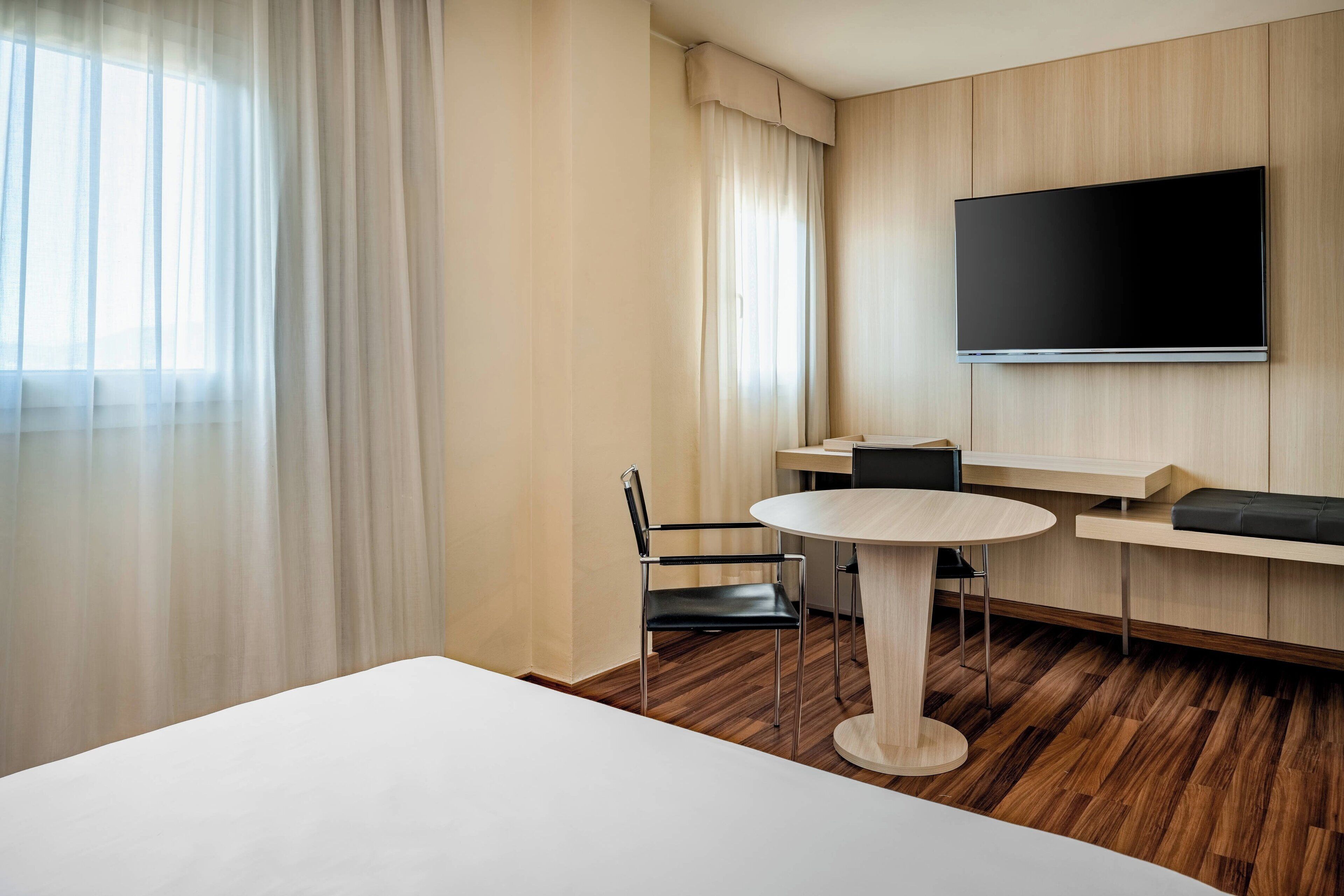 Ein Hotelzimmer mit Bett, rundem Esstisch, Stuhl, an der Wand montiertem Fernseher und Fenster mit leichtem Vorhang.