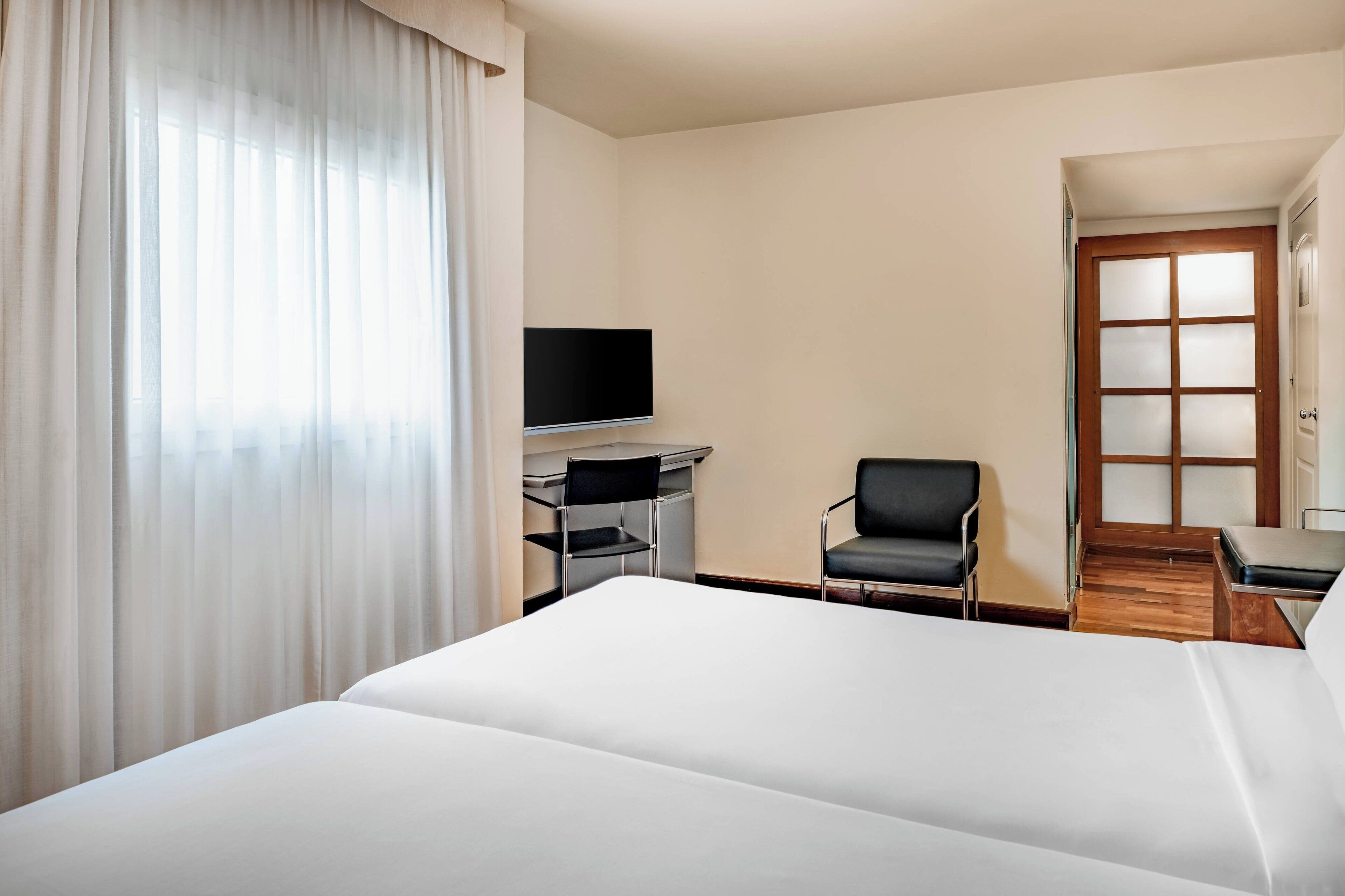 Una habitación de hotel con cama, escritorio con silla, televisor y puerta con paneles de vidrio esmerilado.