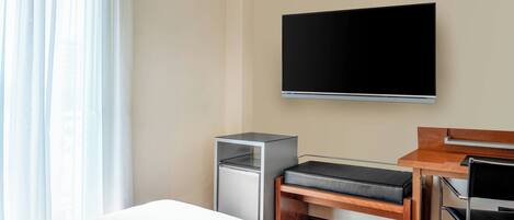 A hotel room with a bed, a desk with a chair, a flat-screen TV, a small refrigerator, and a window with curtains.
