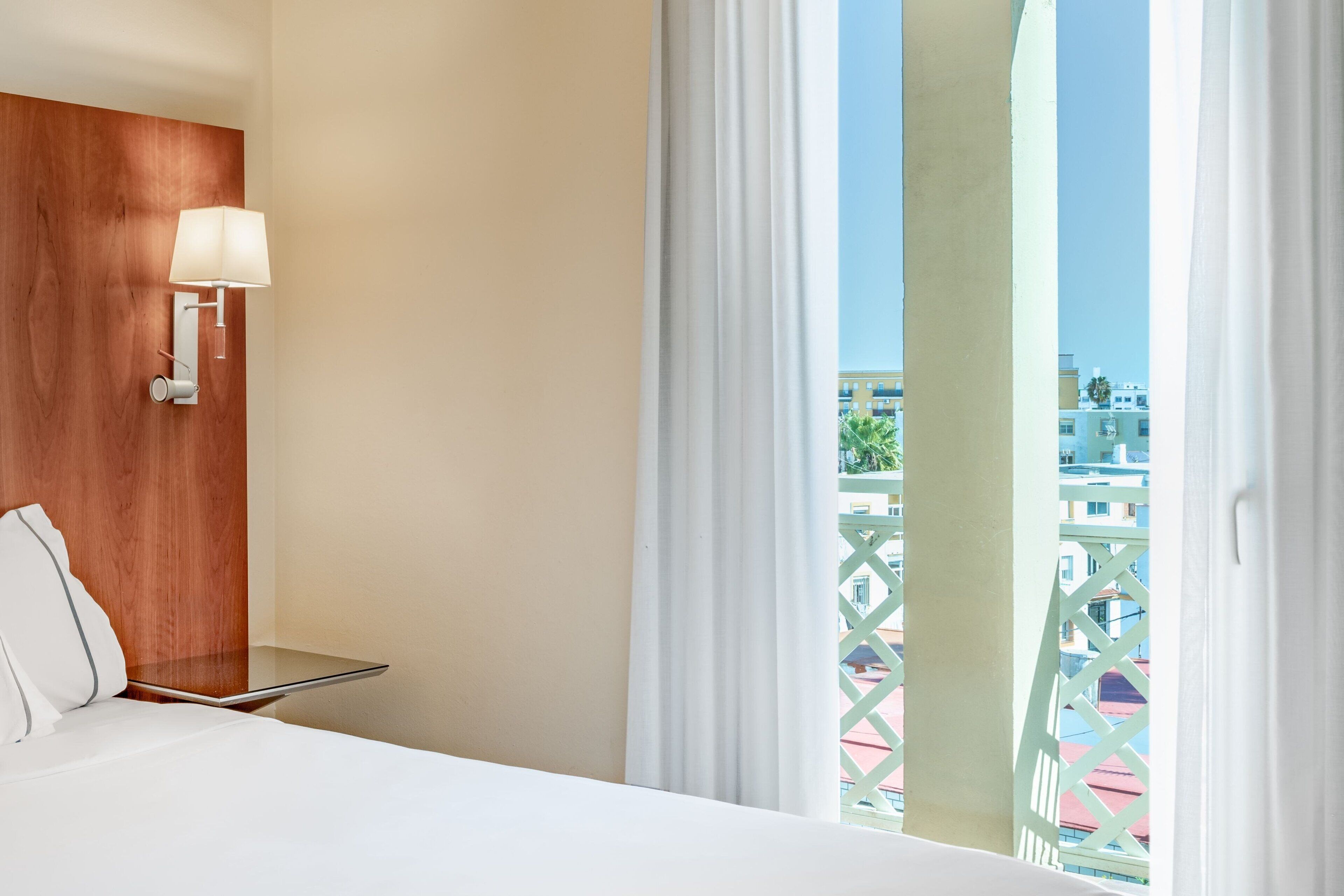A hotel room with a bed, a bedside table, a lamp, and a view of buildings through the window.