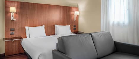 A hotel room with a wooden headboard, two beds with white linens, a grey sofa, and two wall-mounted lamps.