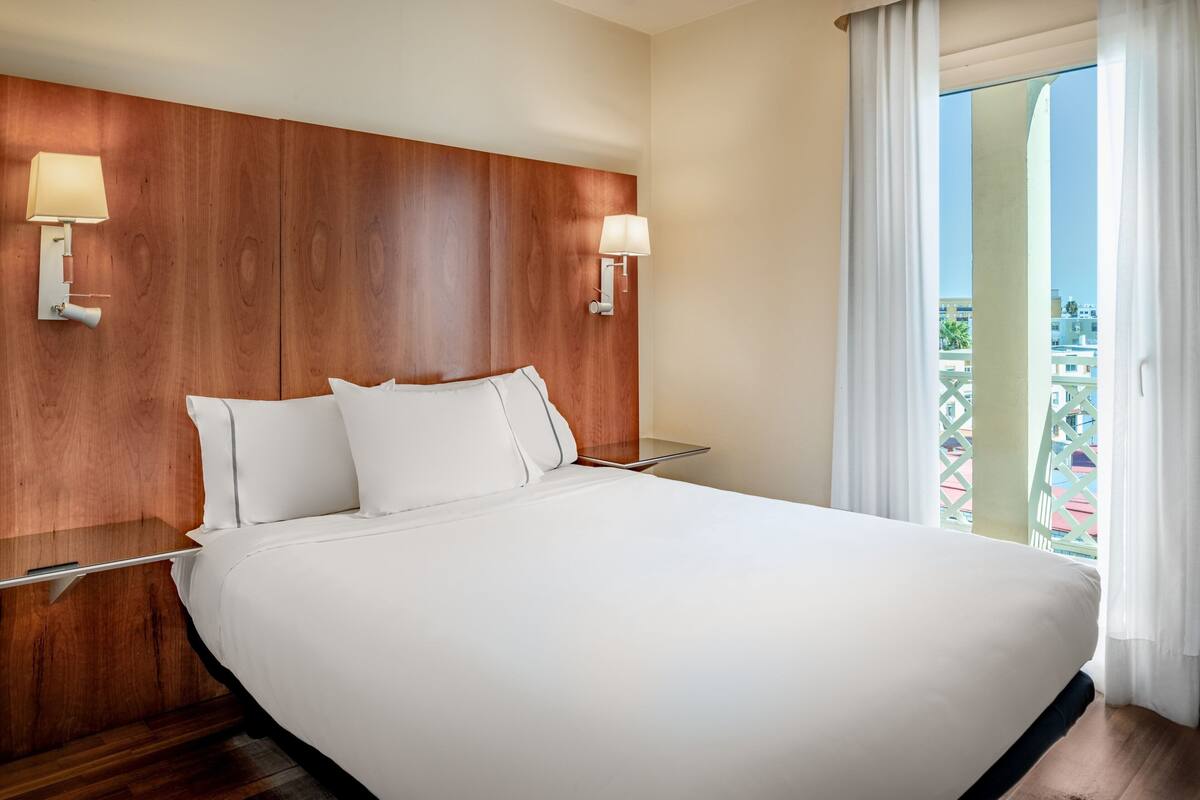 A large bed with white linens, wooden headboard, two bedside lamps, and a view of buildings through a balcony with white curtains.