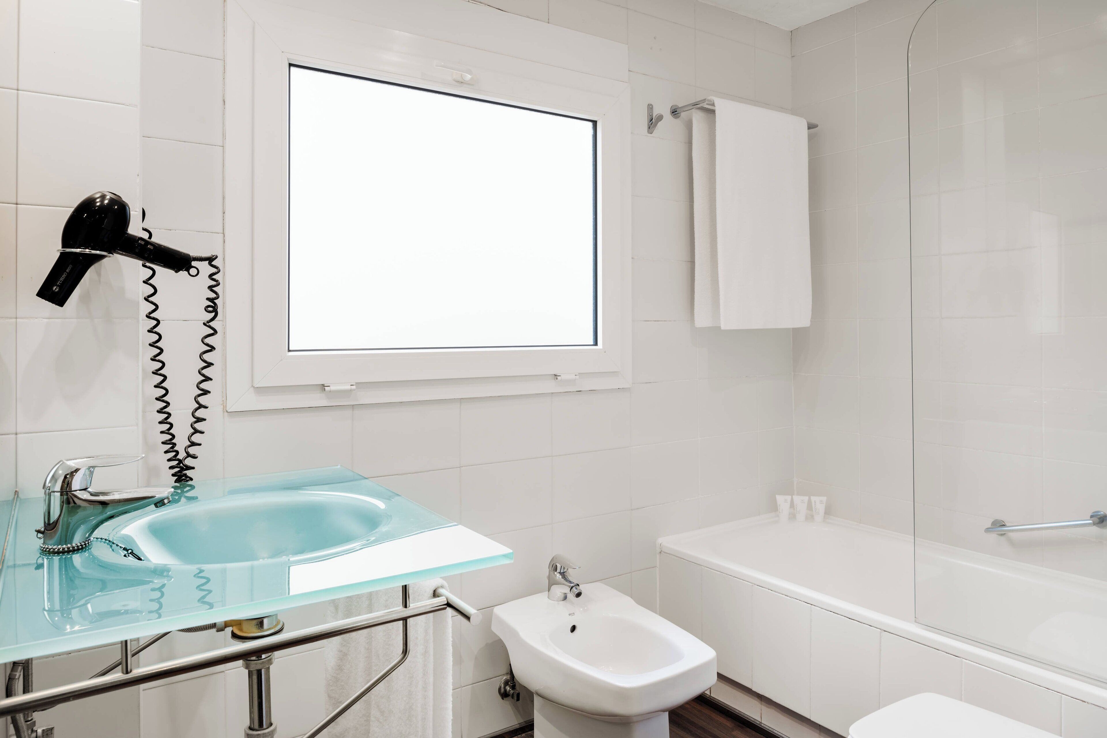 Un baño con lavamanos de vidrio, un bidé, un perchero para toallas y un secador de pelo.